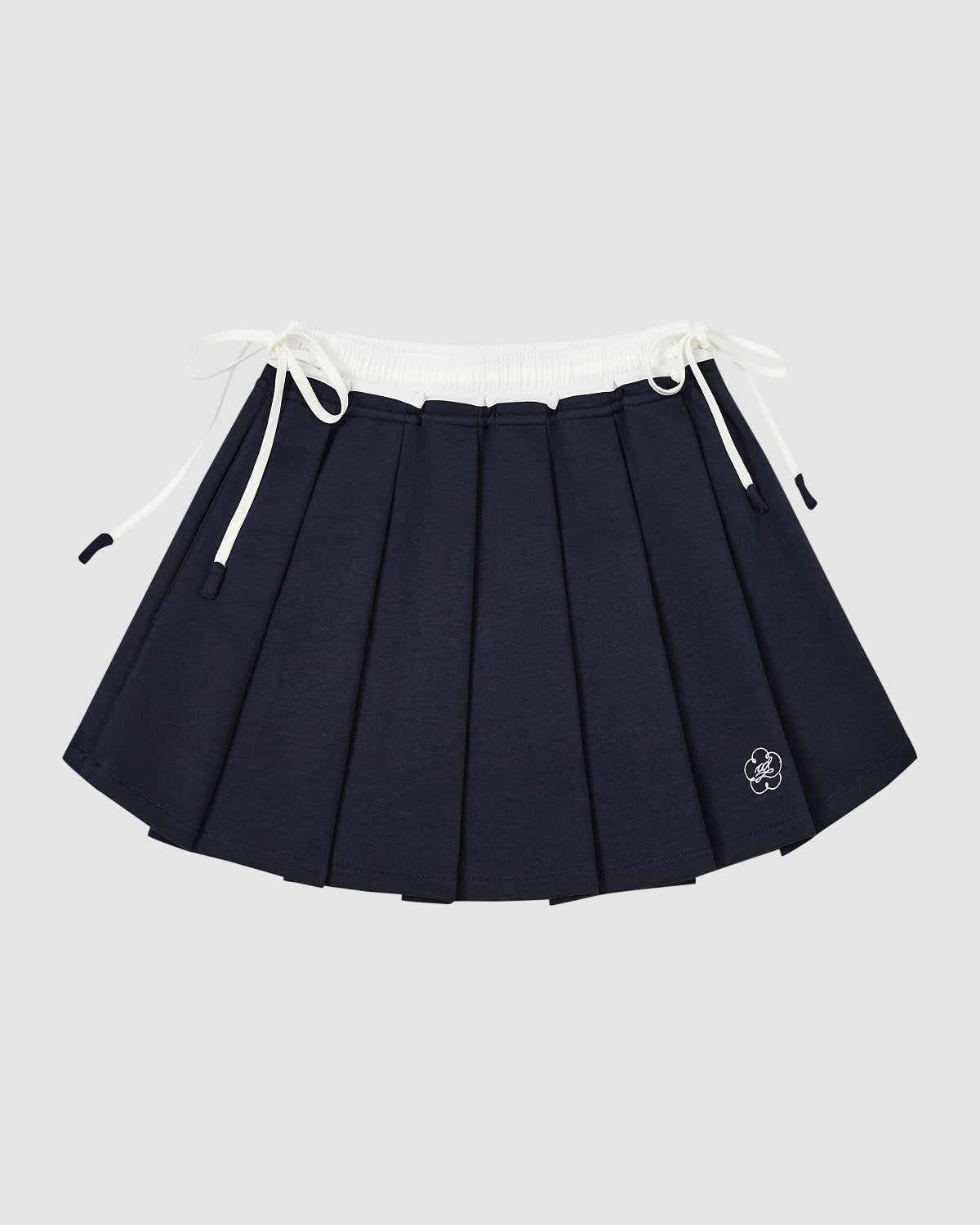 The NAVY PLEATED JERSEY MINI SKIRT by Sundae School features a white elastic waistband, drawstring ties on both sides, and a small embroidered flower near the hem, shown against a plain light gray background.