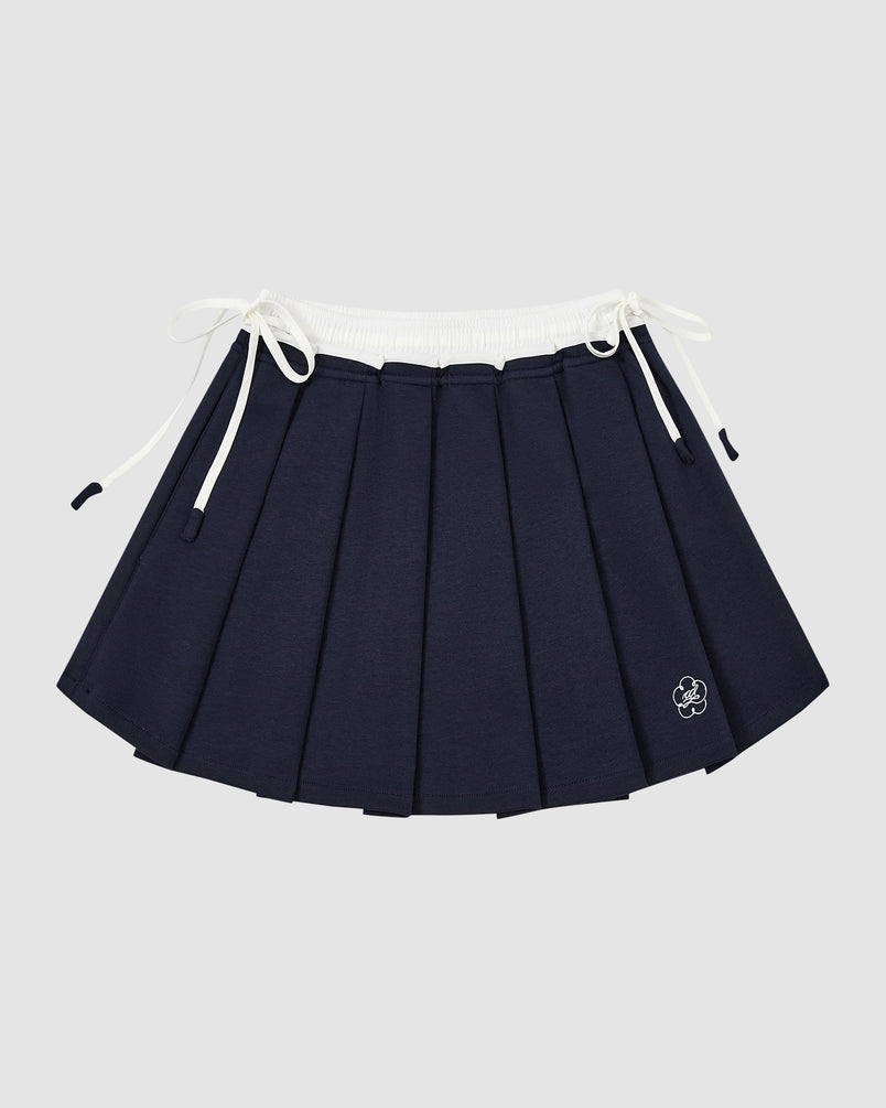 The NAVY PLEATED JERSEY MINI SKIRT by Sundae School features a white elastic waistband, drawstring ties on both sides, and a small embroidered flower near the hem, shown against a plain light gray background.