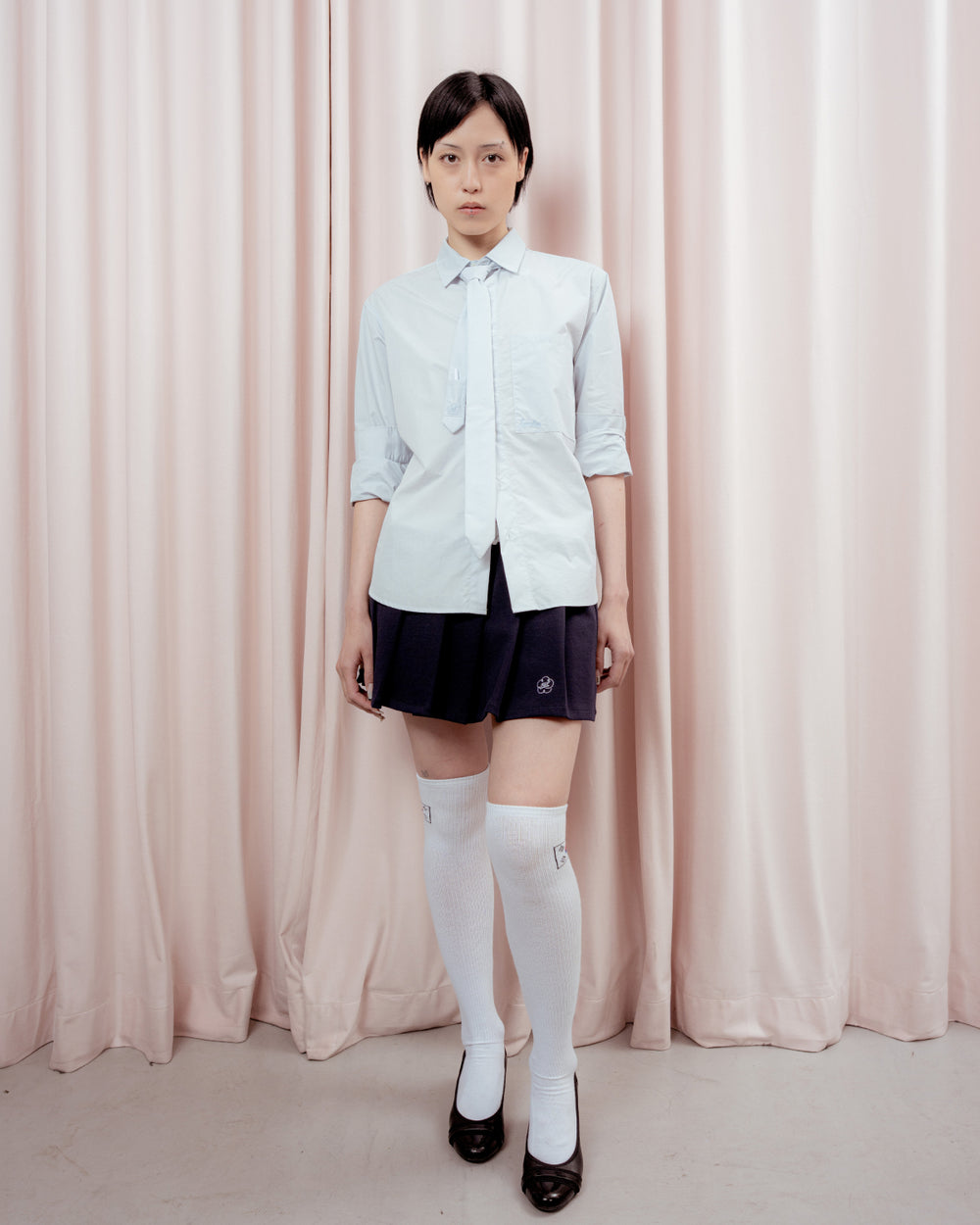 Full-length front view of a person with short black hair standing against a light pink curtain backdrop. They are wearing a light blue button-down shirt with a matching light blue tie, a dark navy pleated mini skirt featuring a small white embroidered logo, white knee-high socks, and black low-heeled shoes.