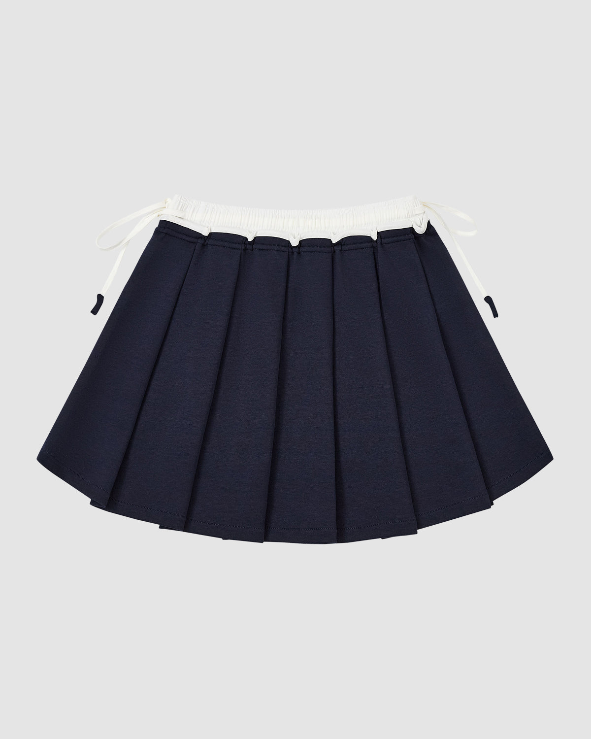 The Sundae School NAVY PLEATED JERSEY MINI SKIRT features a schoolgirl-inspired navy pleated design with a white elastic waistband and drawstrings, shown against a plain light gray background.