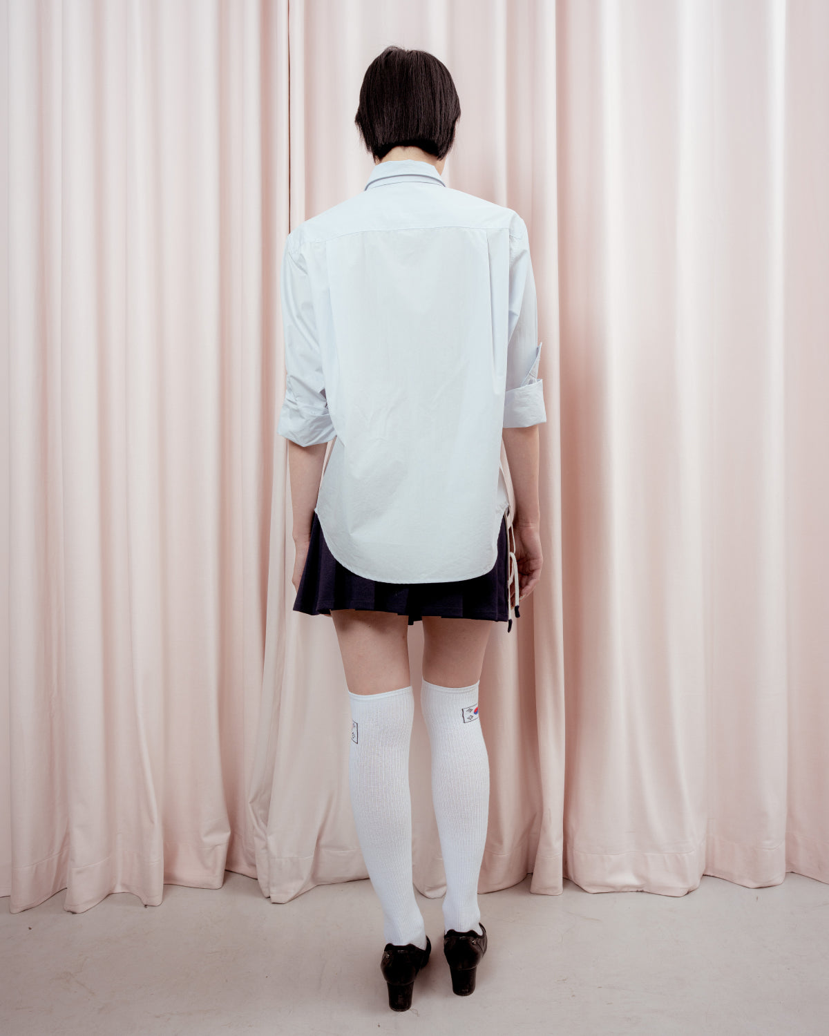 Full-length back view of a person with short black hair standing against a light pink curtain. They are wearing an oversized light blue button-down shirt, a dark navy pleated mini skirt, white knee-high socks with small graphic details, and black low-heeled shoes.