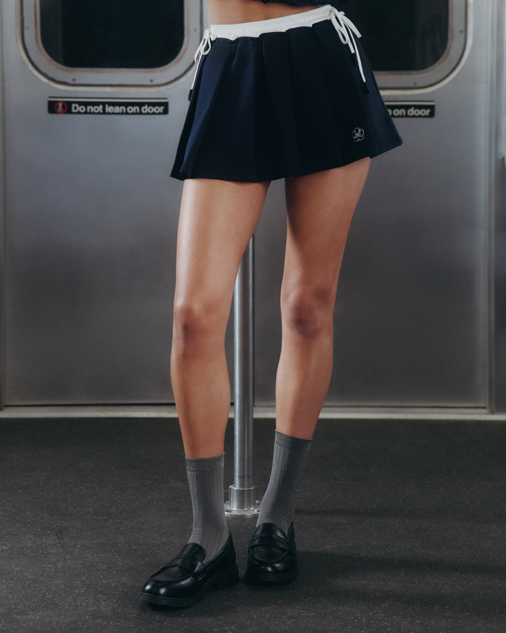 A person stands inside a subway train, wearing Sundae School's NAVY PLEATED JERSEY MINI SKIRT, paired with gray socks and black loafers; their upper body is out of frame.