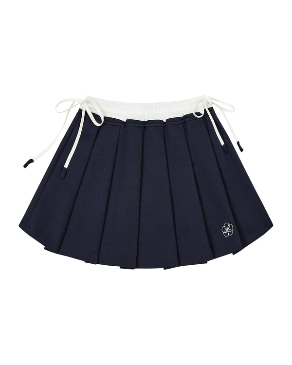 The NAVY PLEATED JERSEY MINI SKIRT by Sundae School features navy blue knife pleats, a white elastic waistband, white drawstrings on each side, and a small embroidered flower near the hem.