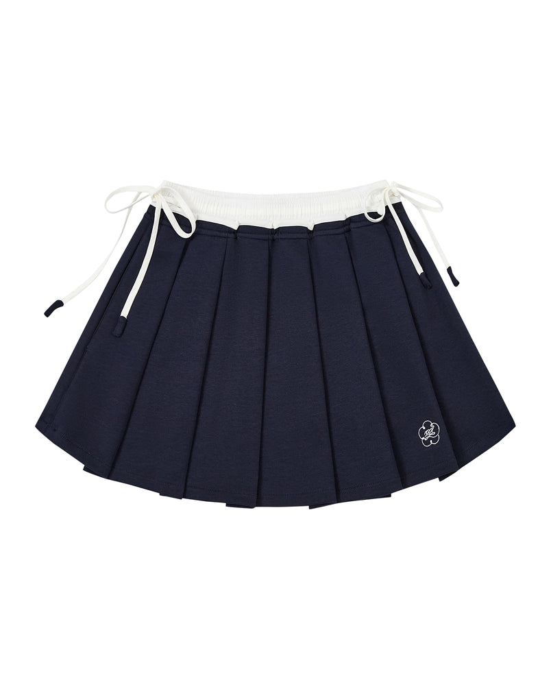 The NAVY PLEATED JERSEY MINI SKIRT by Sundae School features navy blue knife pleats, a white elastic waistband, white drawstrings on each side, and a small embroidered flower near the hem.