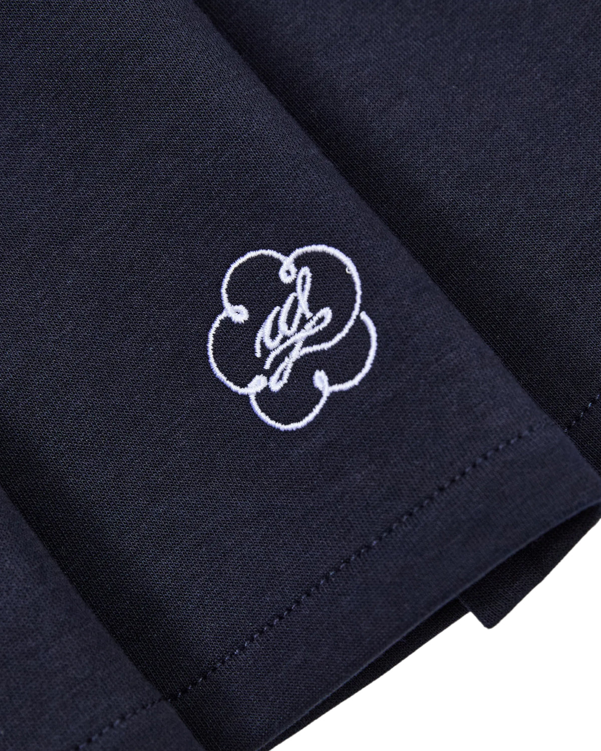 Close-up of the NAVY PLEATED JERSEY MINI SKIRT by Sundae School, crafted from a navy blue cotton blend jersey. The skirt features a white embroidered logo—a stylized W encircled by a looping floral outline. The image highlights the crisp stitching and textured fabric, emphasizing the skirt’s schoolgirl-inspired quality and craftsmanship.