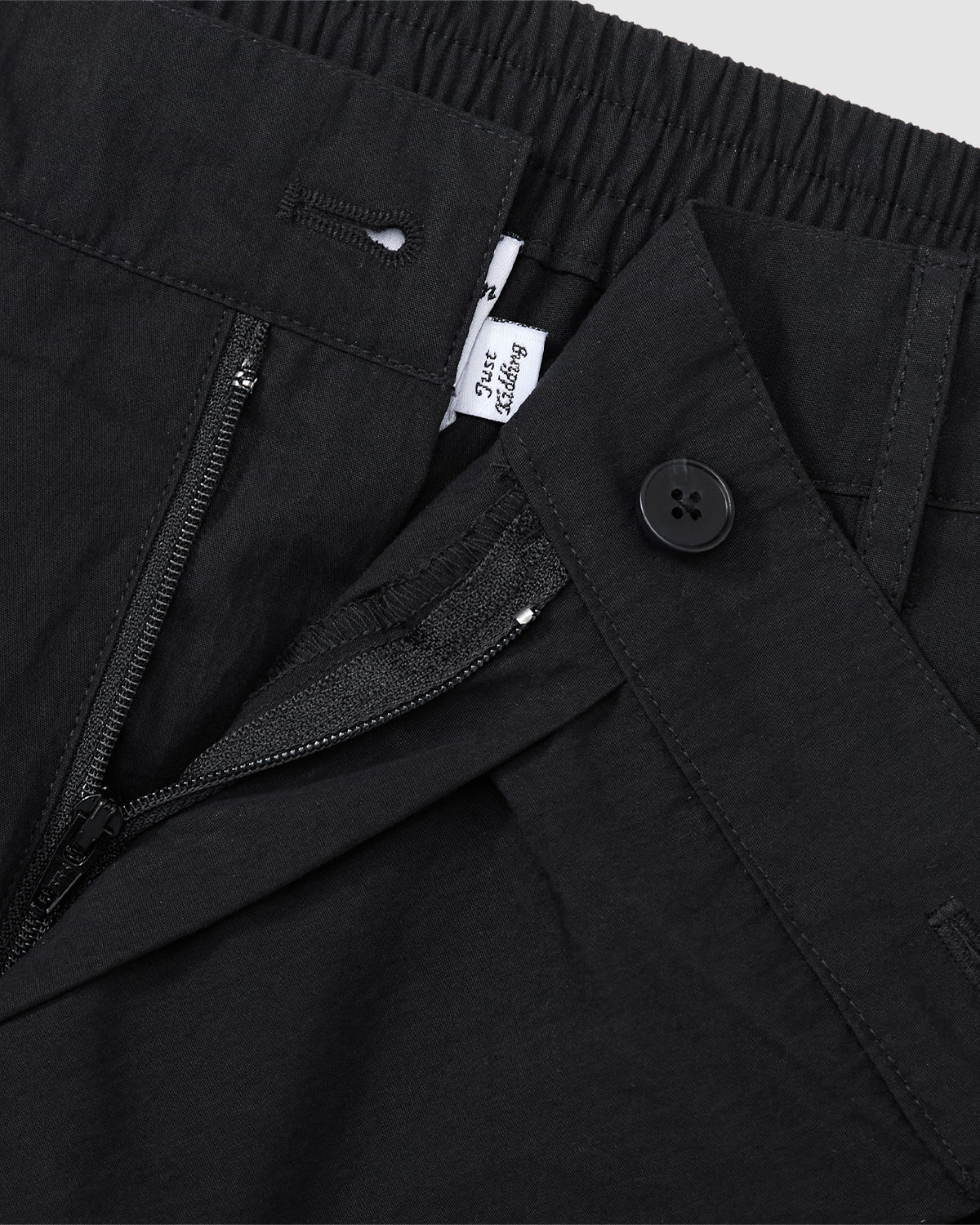 Close-up of Sundae School BLACK PLEATED PALAZZO PANTS with an elastic waistband, zipper fly, black mochi button detail, and a glimpse of a white label featuring black text.