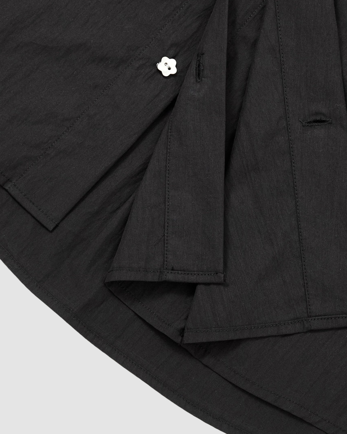 A close-up of the BLACK POPLIN TIE GORUM SHIRT by Sundae School highlights its black fabric hem with overlapping layers, visible seams, and a single white flower-shaped button fastened through a buttonhole.