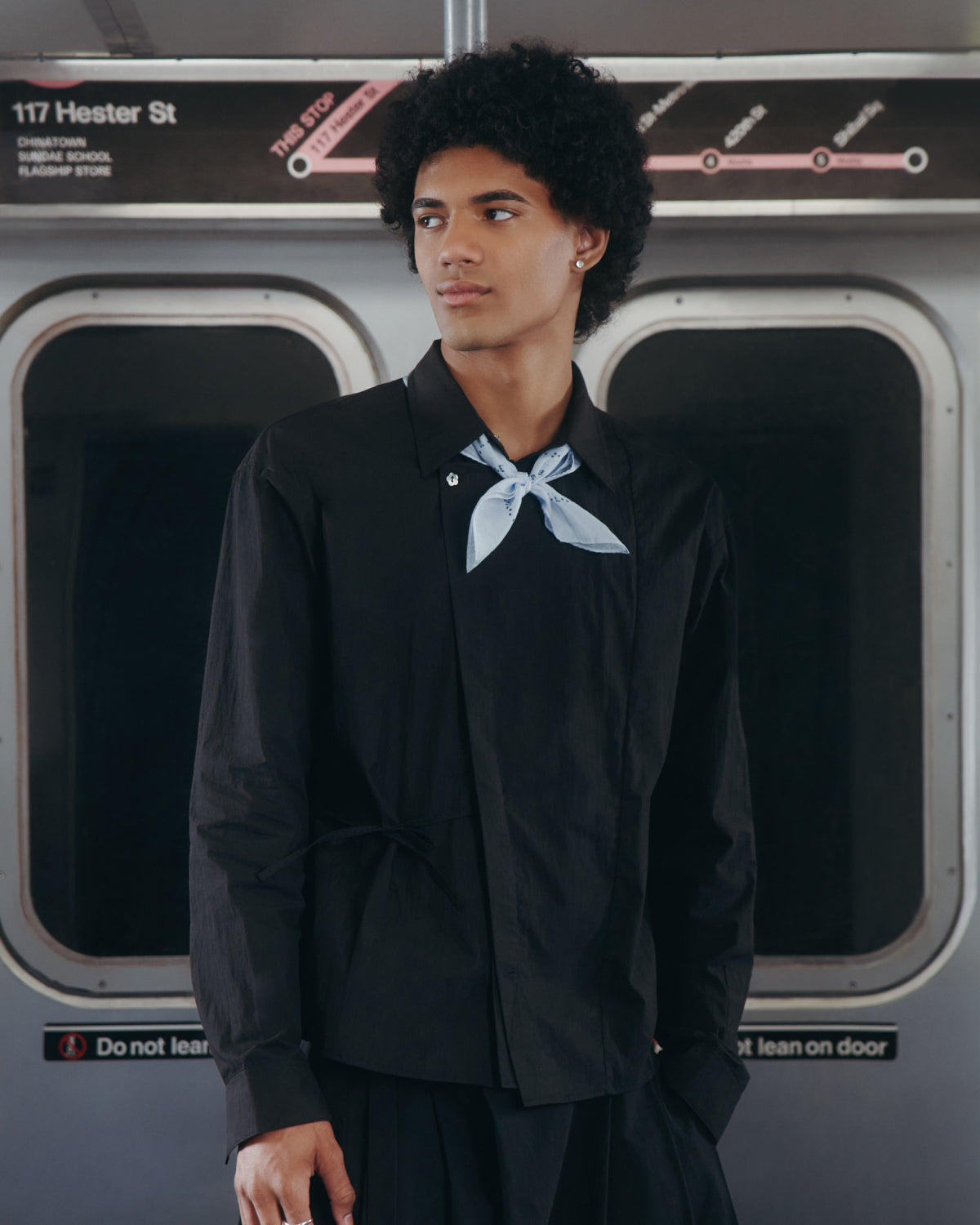 Wearing the BLACK POPLIN TIE GORUM SHIRT by Sundae School and a light blue scarf, a person stands in a subway train in front of a sign that reads 