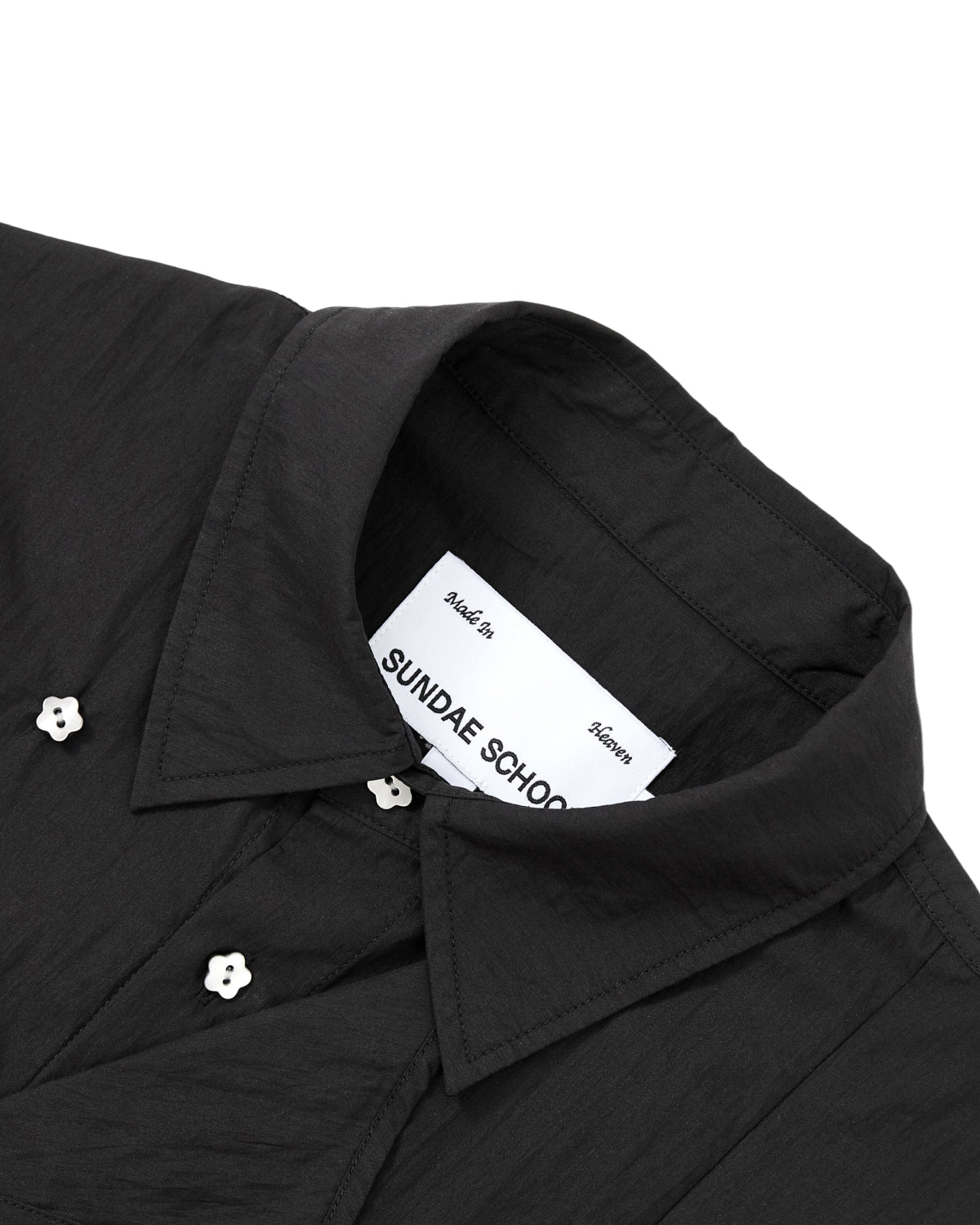 Close-up of the BLACK POPLIN TIE GORUM SHIRT by Sundae School, a unisex tailored-fit black collared shirt crafted from cotton-nylon poplin for a crisp, refined look. The shirt features distinctive white flower-shaped buttons, and the inside label displays the Sundae School branding along with text above the brand name.