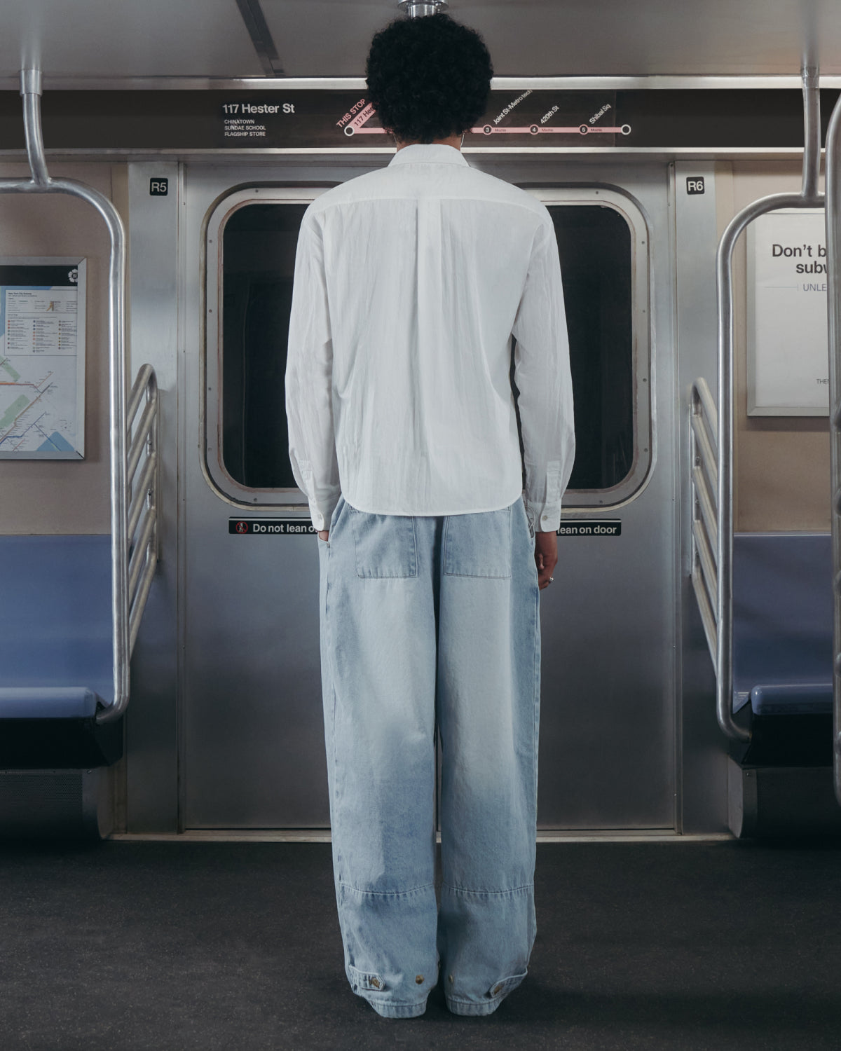Wearing the WHITE POPLIN TIE GORUM SHIRT by Sundae School and baggy light blue jeans, a person with curly hair stands facing the closed doors inside an empty subway car.