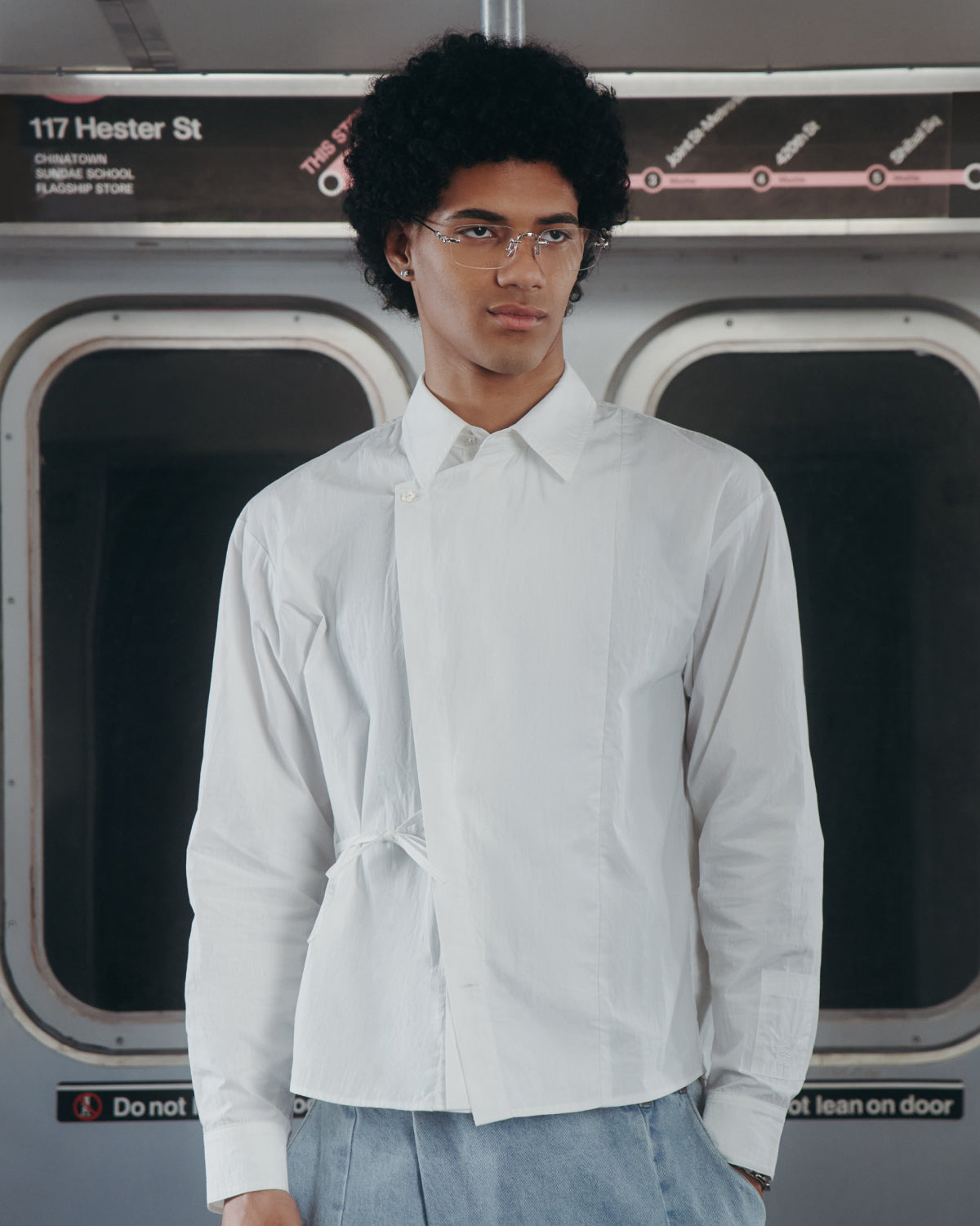 A person with curly hair and glasses wears a Sundae School WHITE POPLIN TIE GORUM SHIRT and stands in a subway train, with windows and a sign that reads 117 Hester St in the background.