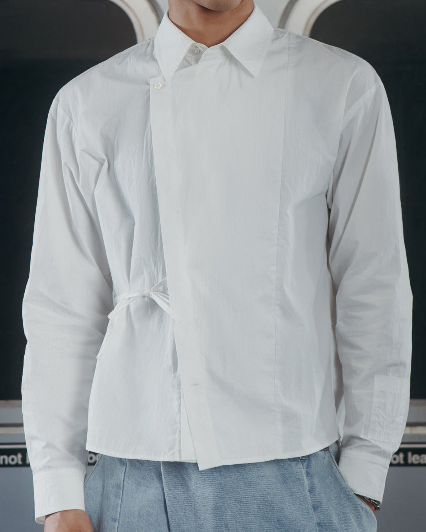 A person with curly hair and glasses wears a Sundae School WHITE POPLIN TIE GORUM SHIRT and stands in a subway train, with windows and a sign that reads 117 Hester St in the background.