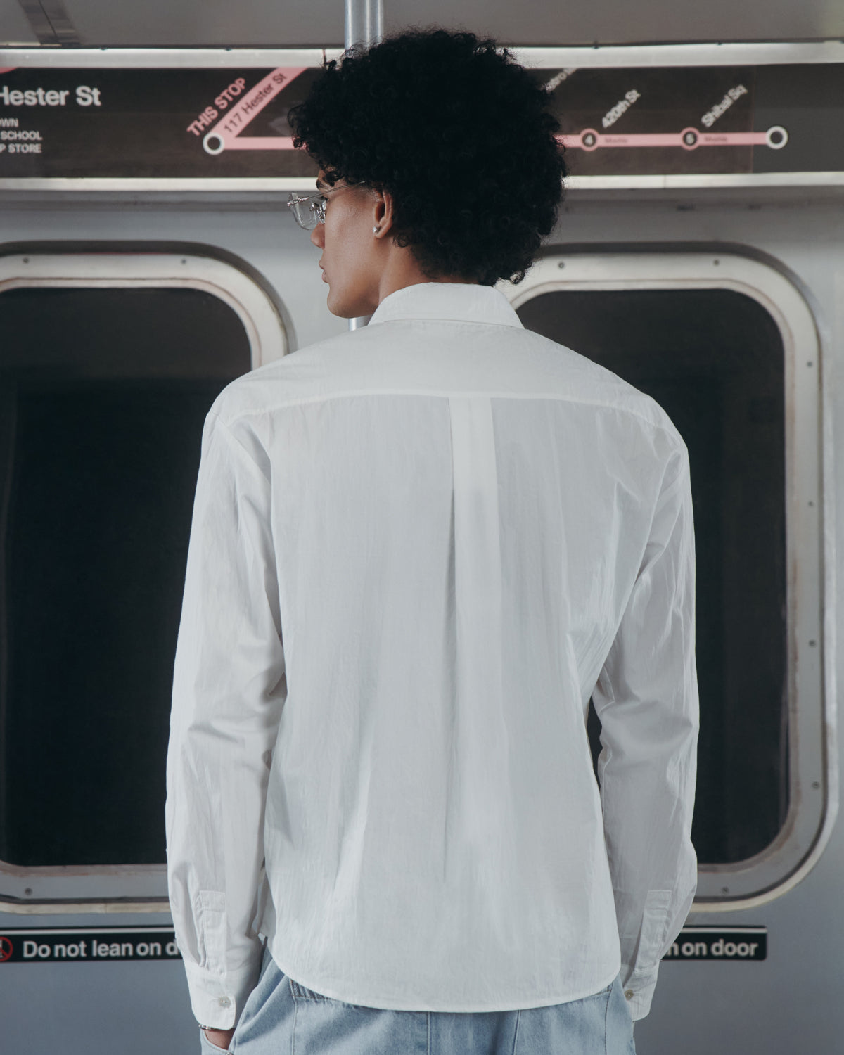 A person with curly hair and glasses, wearing the WHITE POPLIN TIE GORUM SHIRT by Sundae School, stands facing away inside a subway train, with a subway map and station signs visible above the windows in the background.