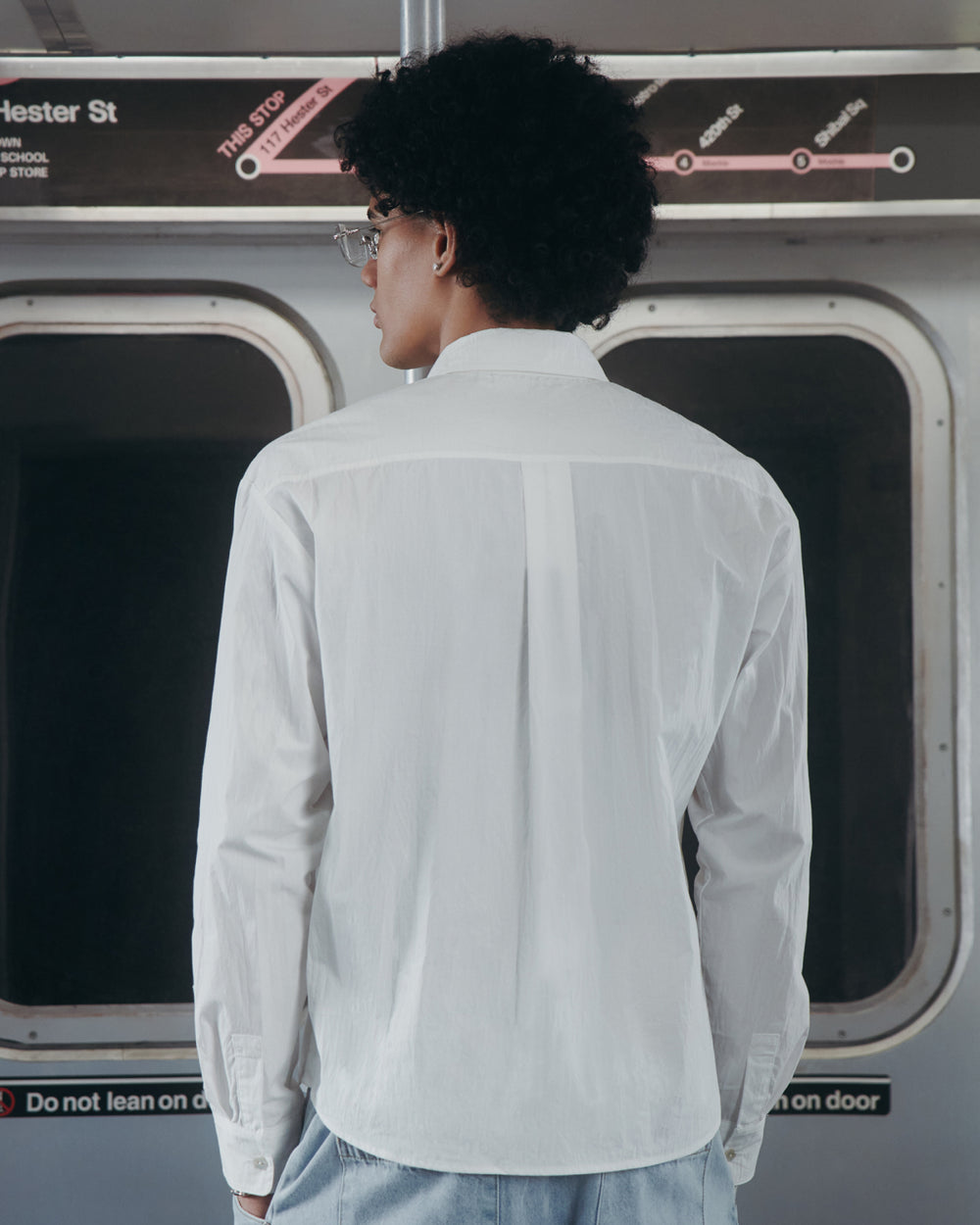 A person with curly hair and glasses, wearing the WHITE POPLIN TIE GORUM SHIRT by Sundae School, stands facing away inside a subway train, with a subway map and station signs visible above the windows in the background.