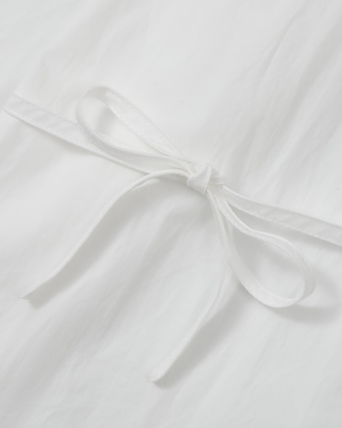 A close-up of a white ribbon tied in a bow on a smooth, white fabric background, evoking the sophisticated detailing of the Sundae School WHITE POPLIN TIE GORUM SHIRT with its signature double-layered front panel.