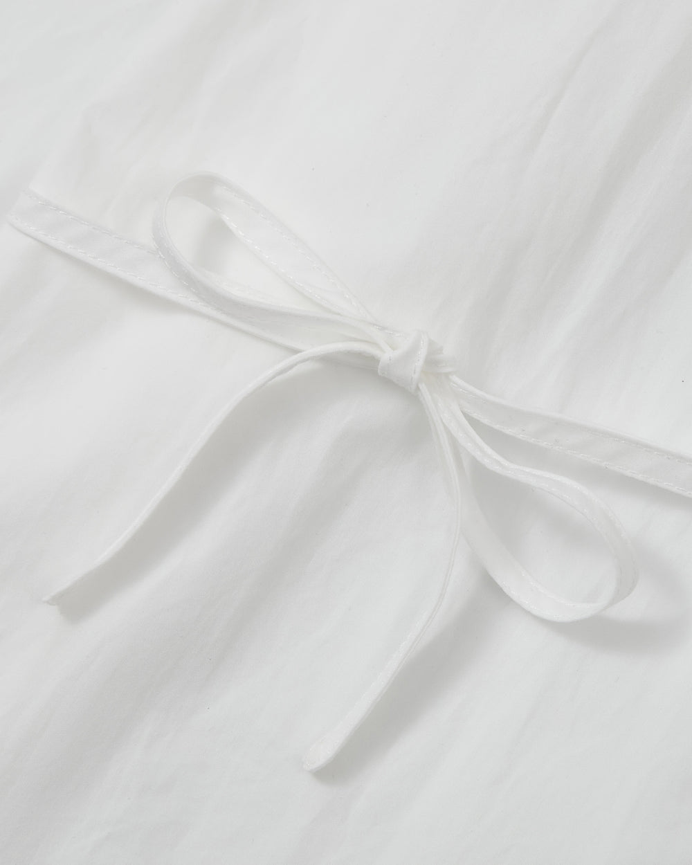 A close-up of a white ribbon tied in a bow on a smooth, white fabric background, evoking the sophisticated detailing of the Sundae School WHITE POPLIN TIE GORUM SHIRT with its signature double-layered front panel.