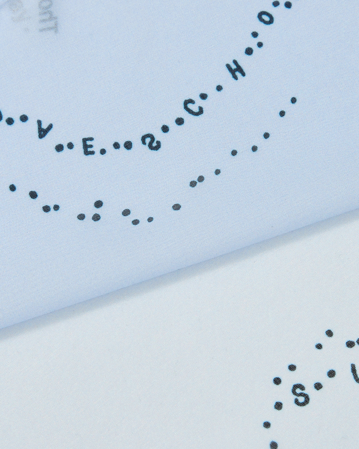 Close-up of a page with black ink letters and dots arranged in a curved line, partially visible on light blue paper. The lettering spells out SCHOOL and other words, evoking the bold printed texture found on the Sundae School SKY BLUE 10 COMMANDMENTS ORGANZA BANDANA.