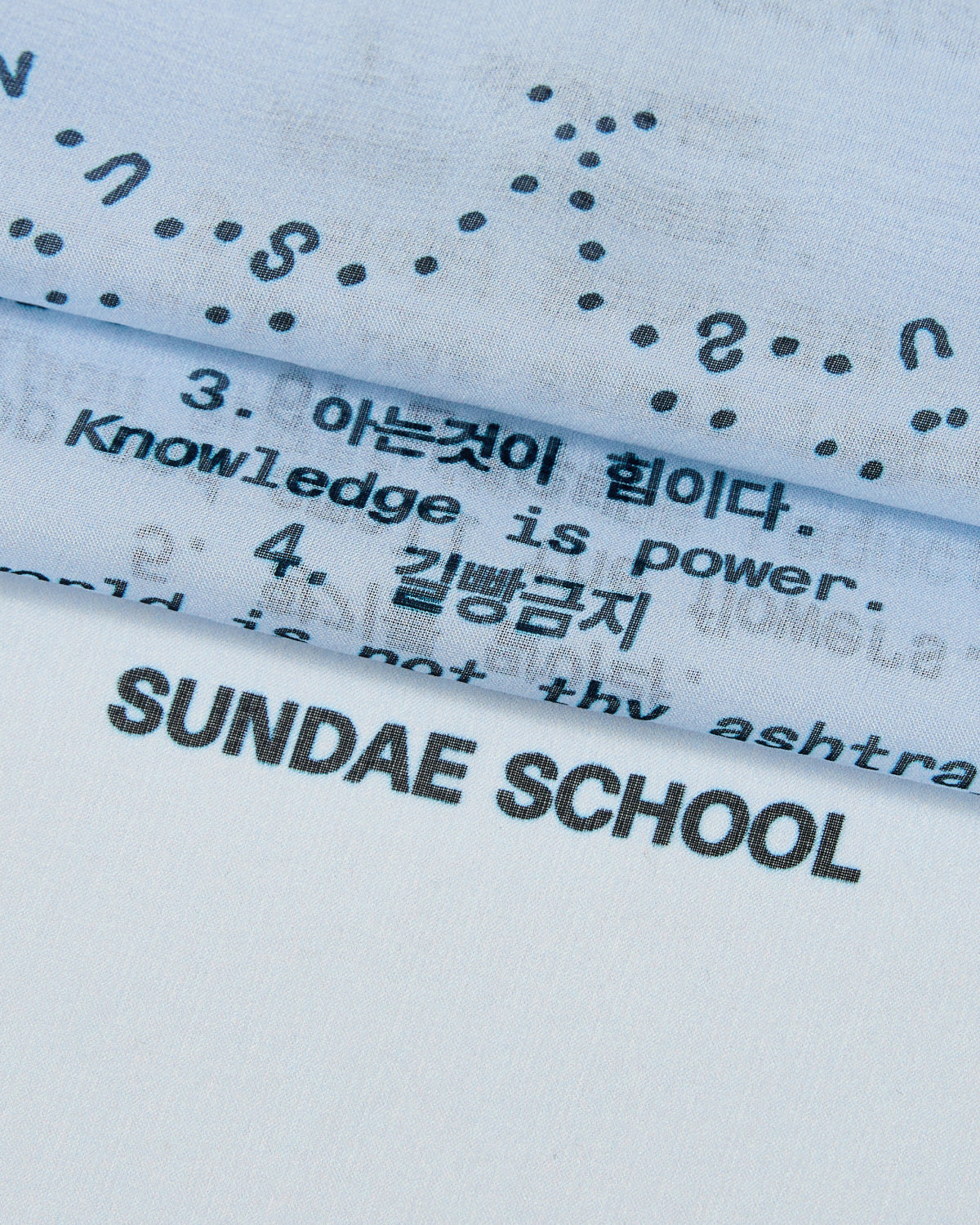 Close-up of the SKY BLUE 10 COMMANDMENTS ORGANZA BANDANA by Sundae School. This sheer silk accessory features black printed text in both English and Korean, including phrases like “Knowledge is power” and “SUNDAE SCHOOL.” The layered organza fabric elegantly showcases the brand’s signature Ten Commandments, resulting in a delicate and stylish look.