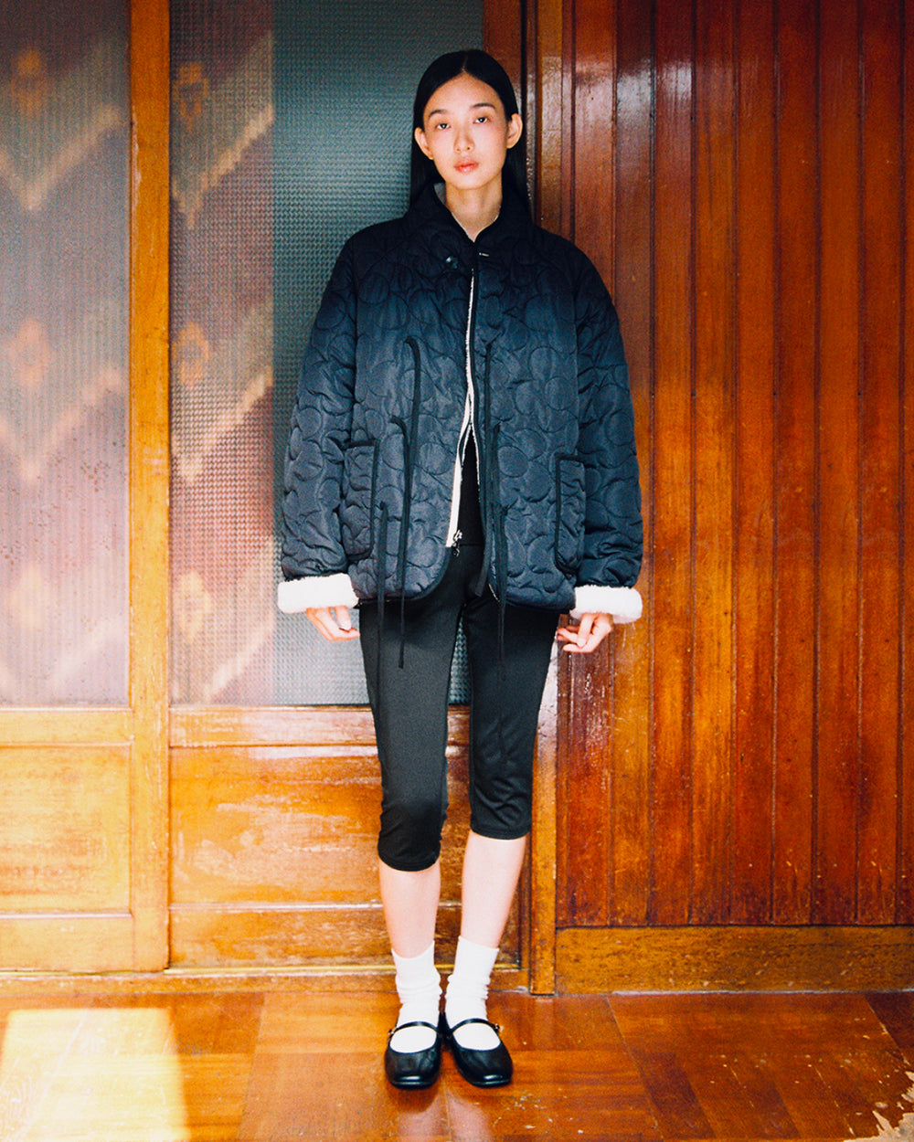 A woman stands indoors against a wooden-paneled background, facing forward with a neutral expression. She is wearing the Sundae School BLACK IVORY REVERSIBLE FLOWER QUILTED JEOGORI FLEECE, paired with black capri leggings, white socks, and black Mary Jane shoes.
