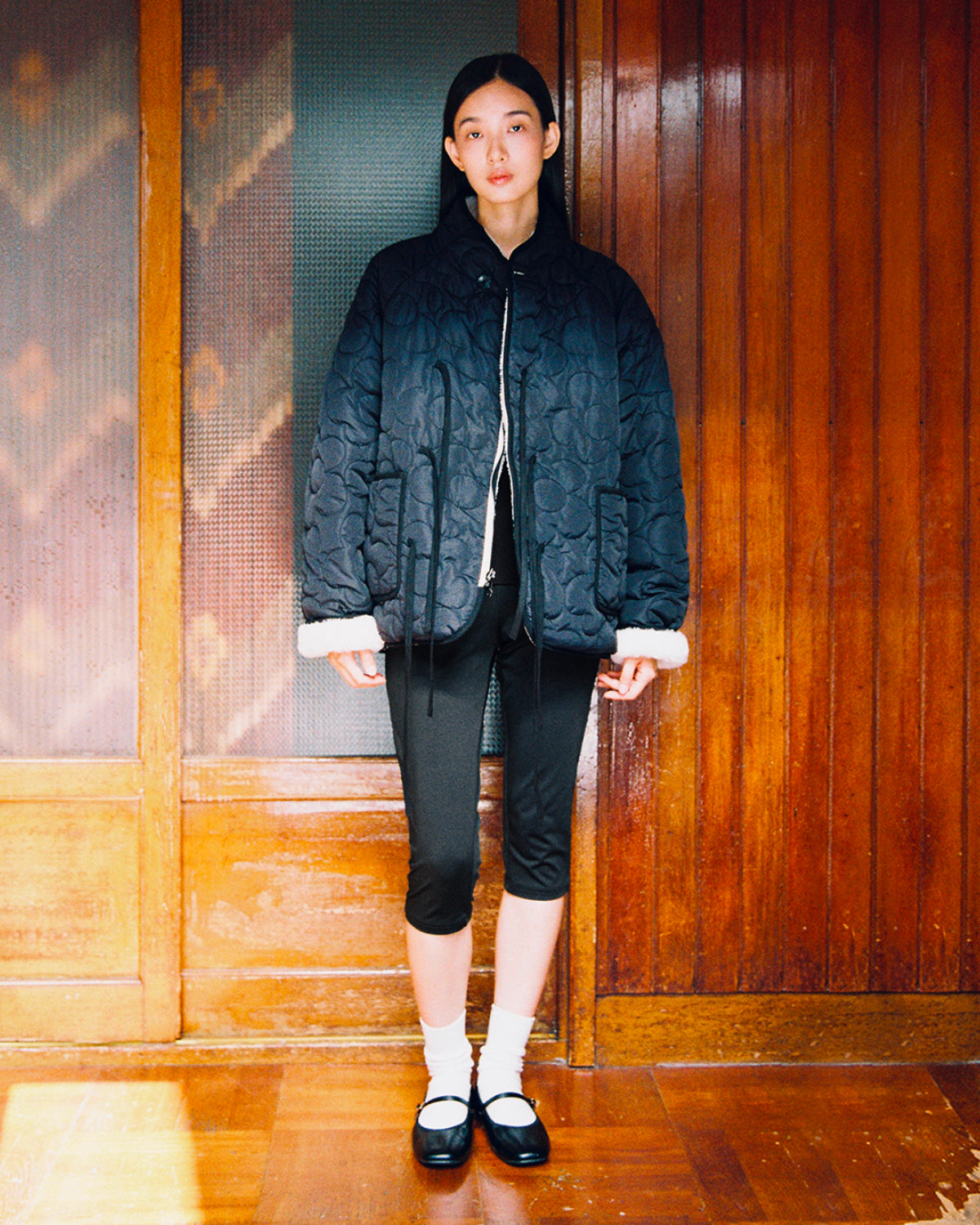 A woman stands indoors against a wooden-paneled background, facing forward with a neutral expression. She is wearing the Sundae School BLACK IVORY REVERSIBLE FLOWER QUILTED JEOGORI FLEECE, paired with black capri leggings, white socks, and black Mary Jane shoes.