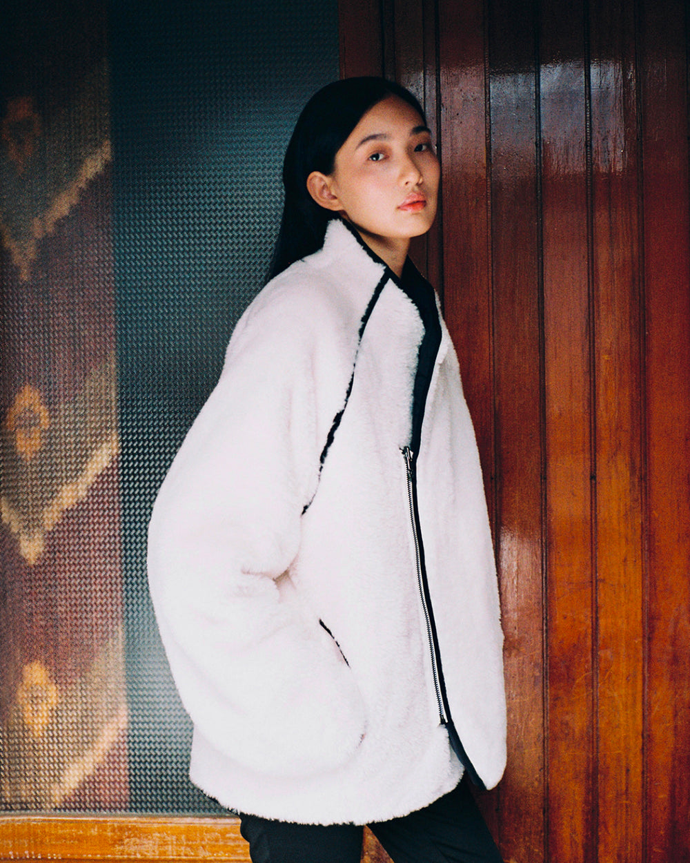Wearing the Sundae School BLACK IVORY REVERSIBLE FLOWER QUILTED JEOGORI FLEECE, a person with straight dark hair stands against a wooden wall and textured glass panel, looking at the camera with their hands in their pockets.