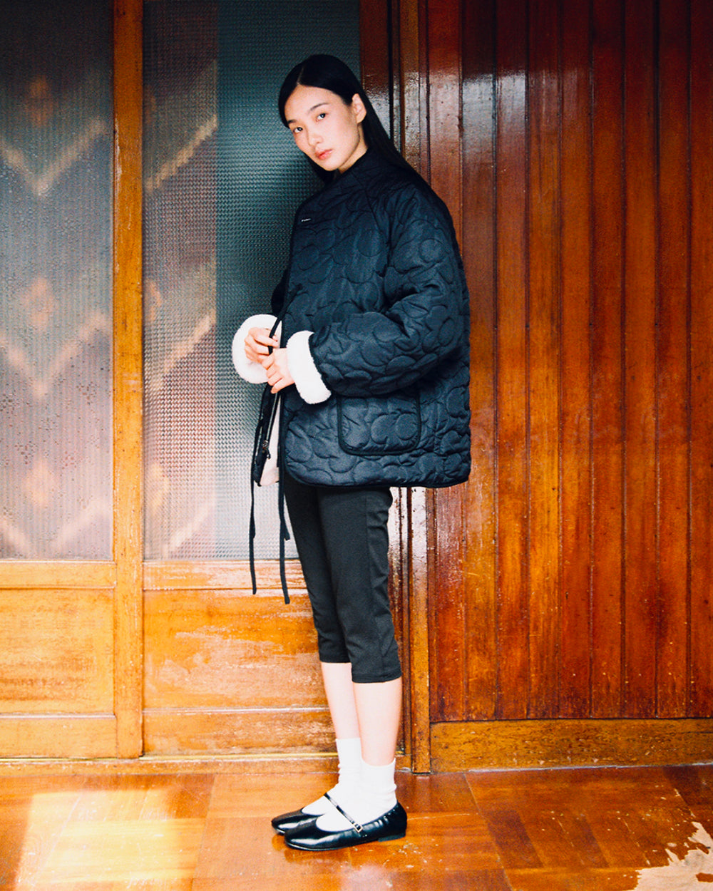 A person stands indoors near a wooden wall and door, facing forward while wearing Sundae School’s BLACK IVORY REVERSIBLE FLOWER QUILTED JEOGORI FLEECE, paired with cropped black pants, white socks, and black Mary Jane shoes.