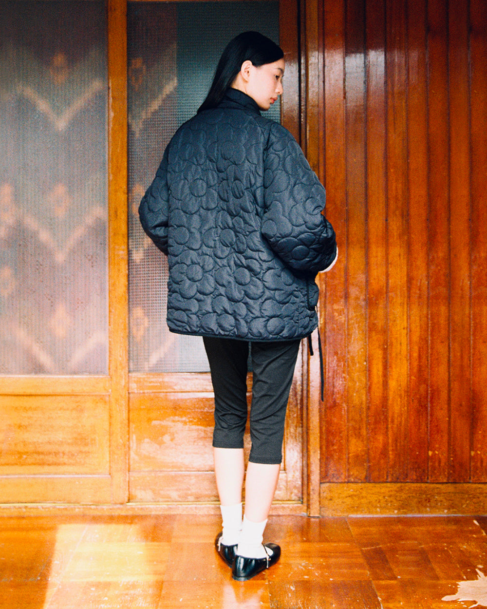 A person stands indoors with their back to the camera, wearing the Sundae School BLACK IVORY REVERSIBLE FLOWER QUILTED JEOGORI FLEECE, paired with cropped black pants, white socks, and black shoes. They are framed by wooden doors and panels.