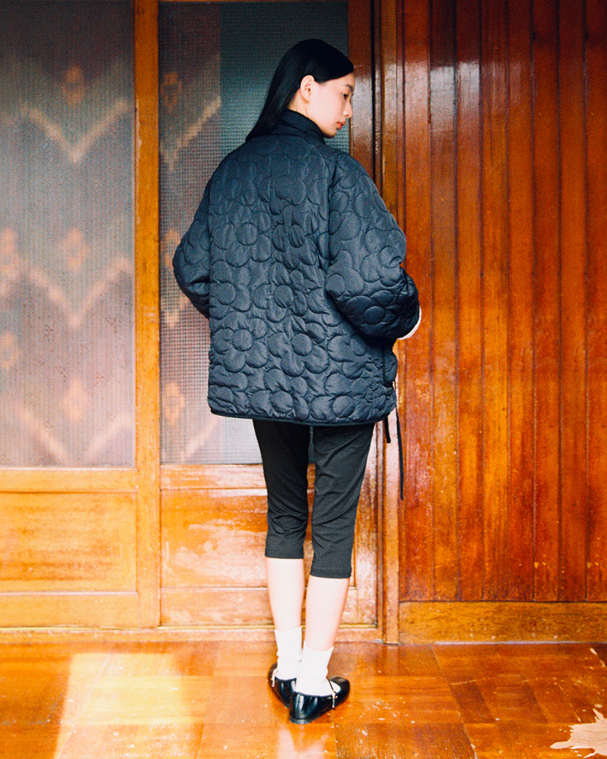 A person stands indoors with their back to the camera, wearing the Sundae School BLACK IVORY REVERSIBLE FLOWER QUILTED JEOGORI FLEECE, paired with cropped black pants, white socks, and black shoes. They are framed by wooden doors and panels.
