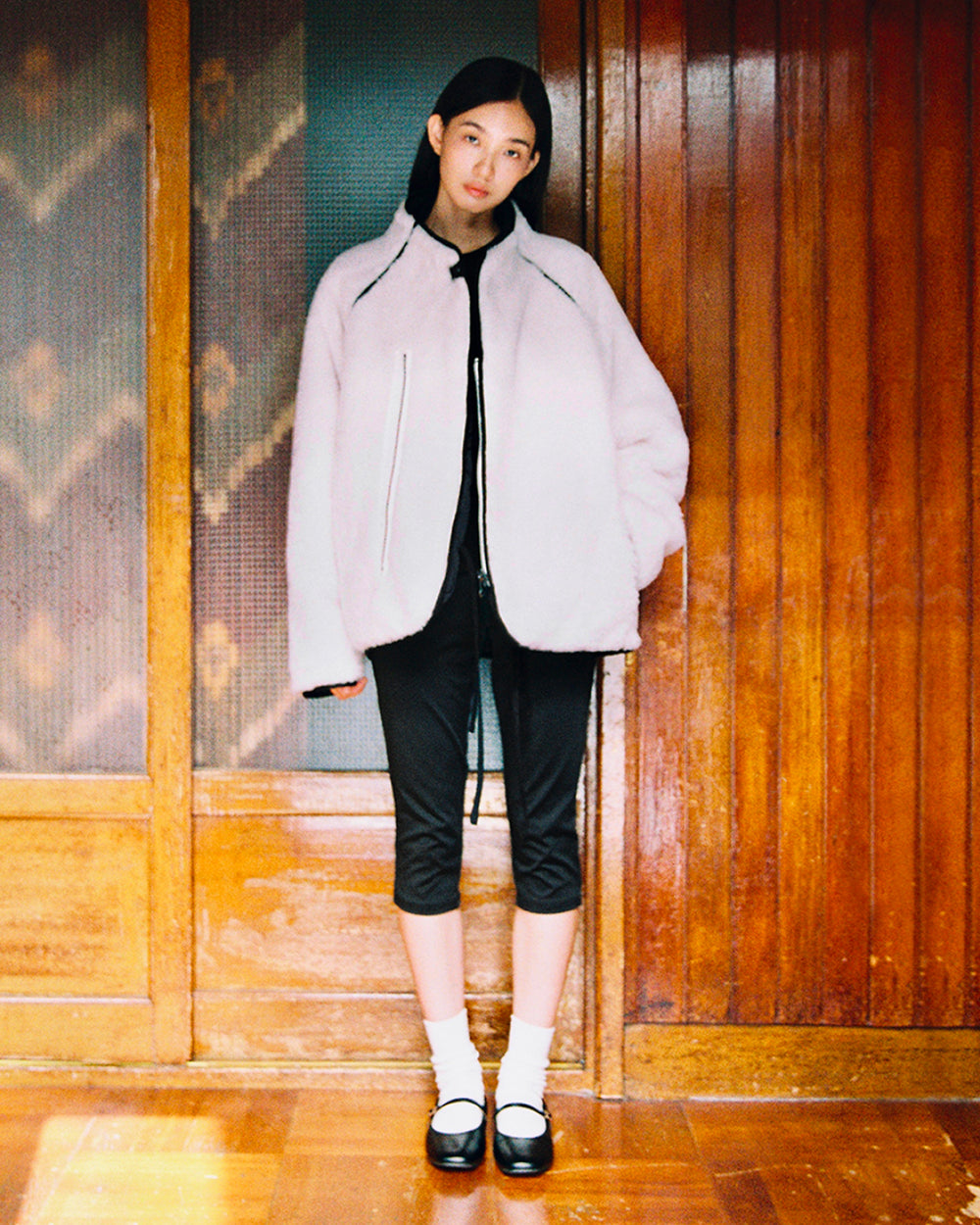 A woman stands indoors against wooden paneling, wearing Sundae School’s BLACK IVORY REVERSIBLE FLOWER QUILTED JEOGORI FLEECE, black cropped pants, white socks, and black Mary Jane shoes. She looks at the camera with a neutral expression.