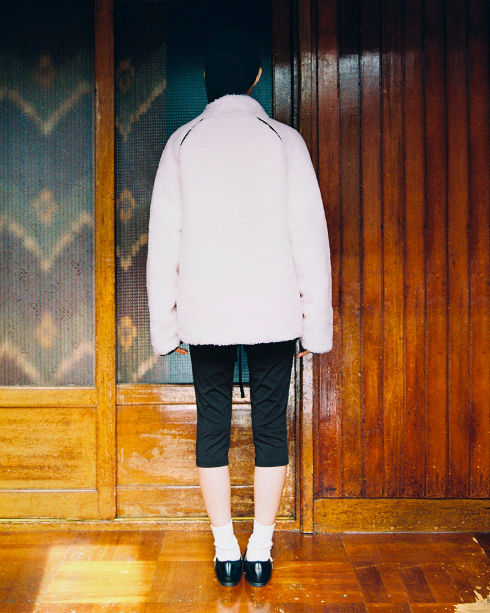 A person stands with their back to the camera on a polished wooden floor, facing a wooden wall. They are wearing the Sundae School BLACK IVORY REVERSIBLE FLOWER QUILTED JEOGORI FLEECE, paired with black pants, white socks, black shoes, and a black head covering.