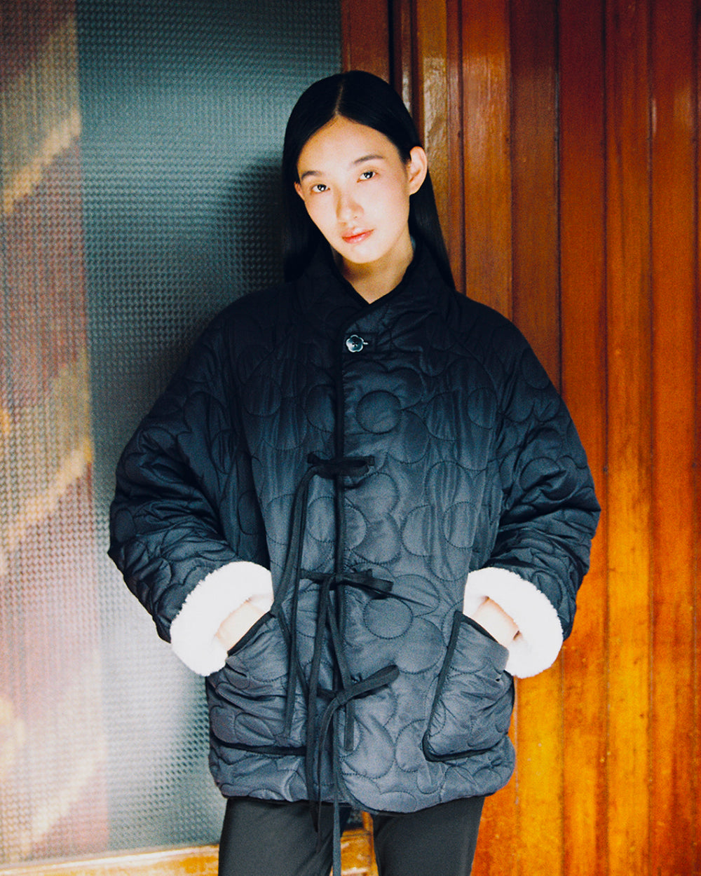 A person with long black hair wears the BLACK IVORY REVERSIBLE FLOWER QUILTED JEOGORI FLEECE by Sundae School, featuring white cuffs, while standing indoors against a wooden wall and glass panel.