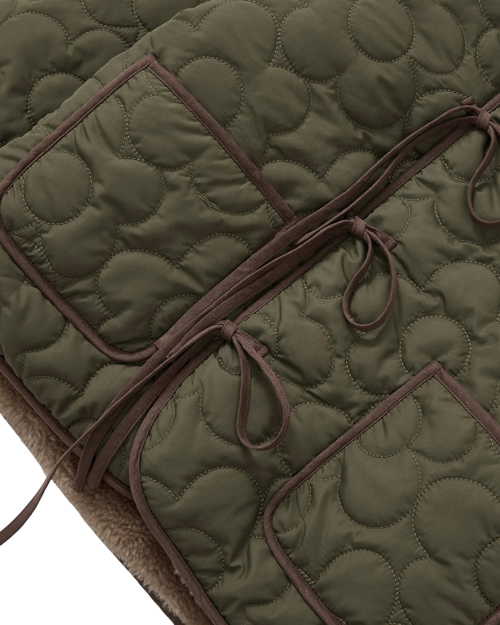 Close-up of the Sundae School KHAKI REVERSIBLE FLOWER QUILTED JEOGORI FLEECE, featuring olive green quilted fabric adorned with circular stitching patterns, two front pockets, brown trim, and tied fabric bows. A peek of soft fleece lining at the edge highlights its hanbok-inspired design and evokes the cozy aesthetic of a faux-sherpa puffer.
