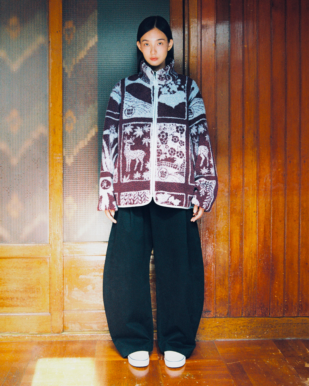 A person stands indoors against a wood-paneled wall, wearing the Sundae School PLUM REVERSIBLE GOSTOP FLEECE—a stand collar zip-up jacket featuring animal and plant designs—paired with wide black pants and white platform shoes.