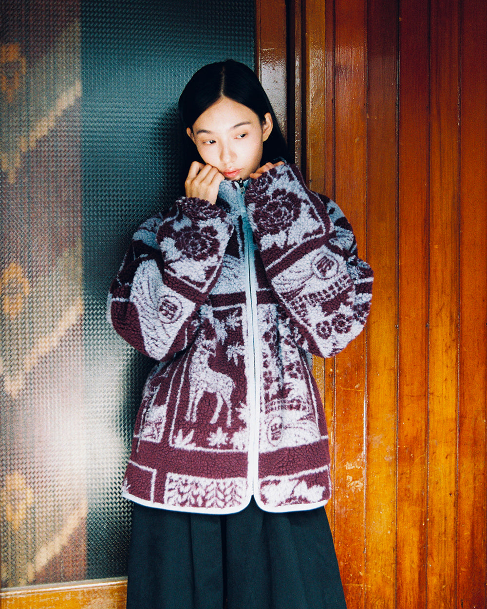A person stands indoors in front of wooden paneling and textured glass, wearing the Sundae School PLUM REVERSIBLE GOSTOP FLEECE—a patterned hwatu jacket adorned with animals and nature motifs—looking down and gently holding the stand collar zip-up.