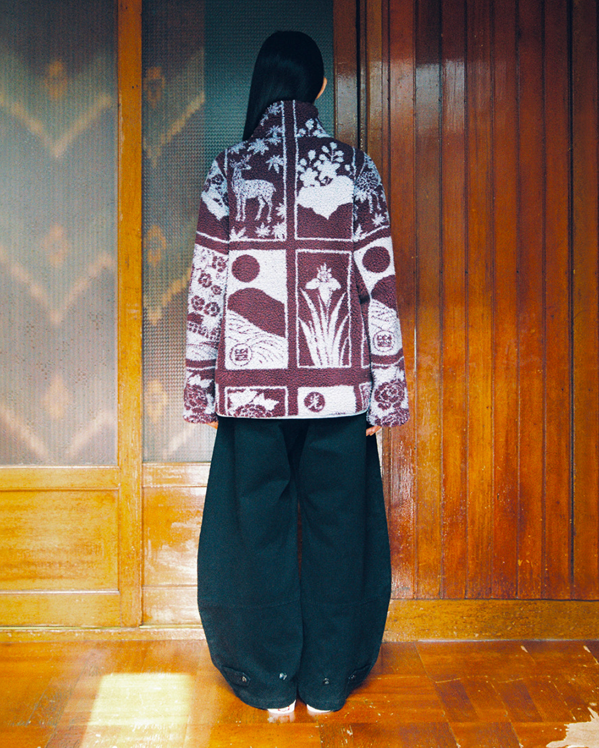 A person stands indoors facing a wooden wall, wearing wide black pants and the Sundae School PLUM REVERSIBLE GOSTOP FLEECE—a patterned purple and white jacket featuring nature-inspired designs and a stand collar zip-up style. Their long, straight dark hair is worn loose down their back.
