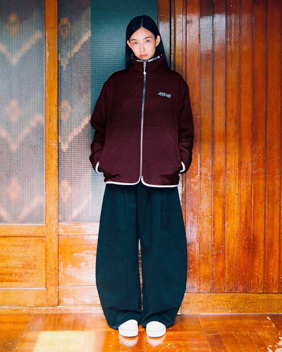 A person stands indoors by wooden walls with hands in their pockets and a neutral expression, wearing Sundae School’s PLUM REVERSIBLE GOSTOP FLEECE in maroon, paired with wide black pants and white platform shoes.