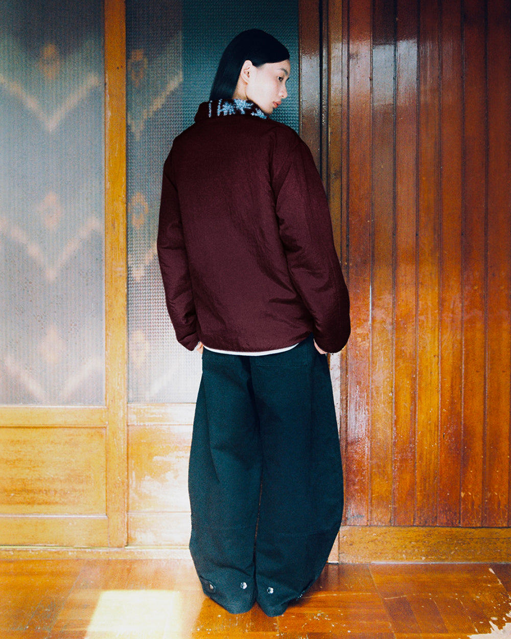 A person with straight black hair stands indoors, facing away, wearing Sundae School’s PLUM REVERSIBLE GOSTOP FLEECE in maroon paired with wide black pants. The reversible sherpa fleece adds both warmth and texture to their look as they stand before wooden panel doors, their head turned slightly to the side.