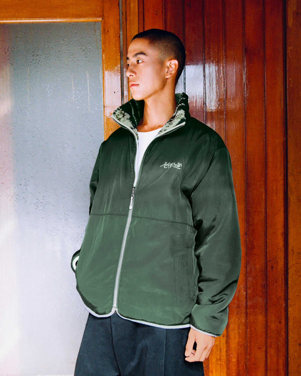 A person with short hair stands indoors against wooden paneling, wearing the Sundae School TEAL REVERSIBLE GOSTOP FLEECE jacket with a sherpa fleece lining over a white t-shirt, looking to the side.