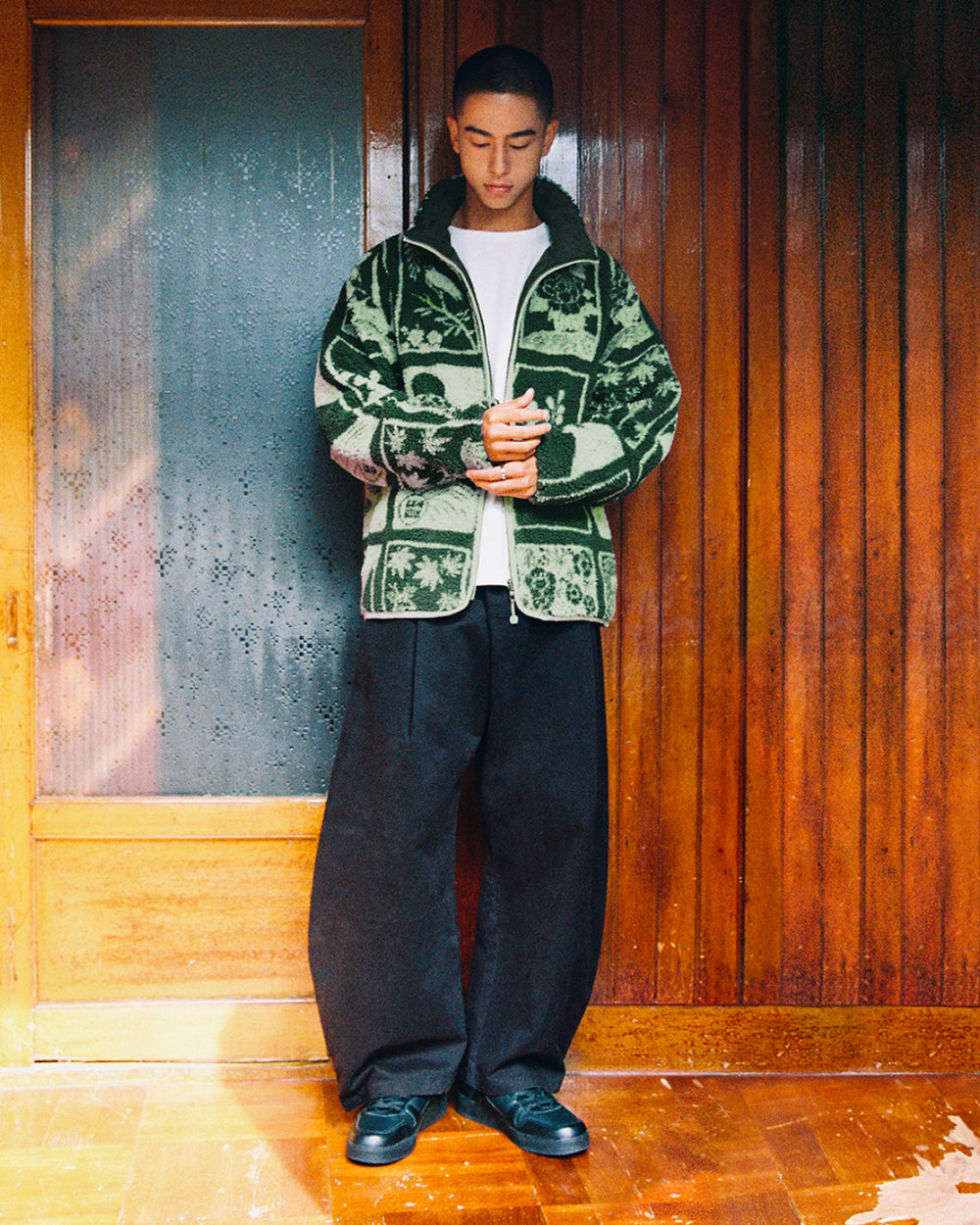 A person stands indoors in front of a wooden door and paneling, wearing a white t-shirt, black wide-leg pants, black sneakers, and the Sundae School TEAL REVERSIBLE GOSTOP FLEECE—a green patterned jacket with a stand collar zip up—with hands clasped and head slightly bowed.