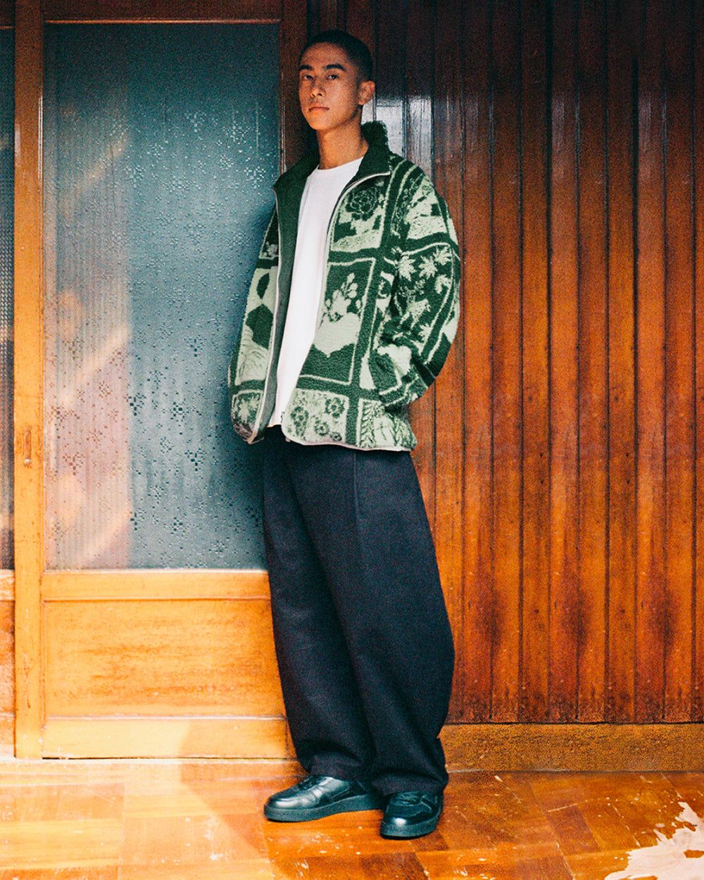 A person stands indoors by a wooden wall, wearing the Sundae School TEAL REVERSIBLE GOSTOP FLEECE with a stand collar zip up over a white shirt, loose black pants, and black shoes, looking at the camera.