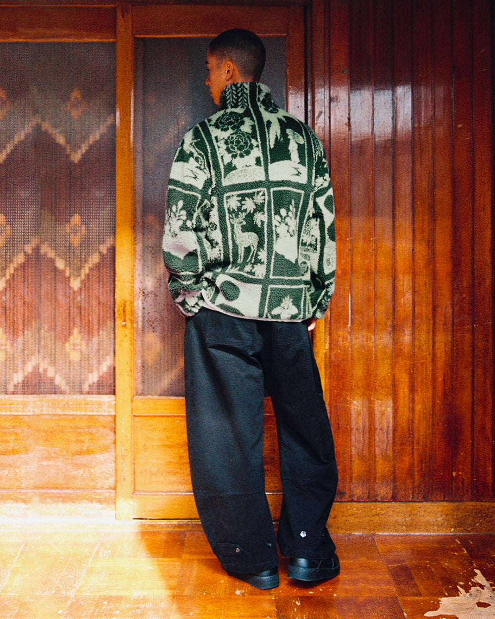 A person stands indoors facing away, wearing the Sundae School TEAL REVERSIBLE GOSTOP FLEECE—a green and white patterned hwatu jacket featuring animal and nature imagery—with loose black pants and black shoes. The reversible sherpa fleece features a stand collar zip-up design for both warmth and style. Wooden paneling and sliding doors are visible in the background.