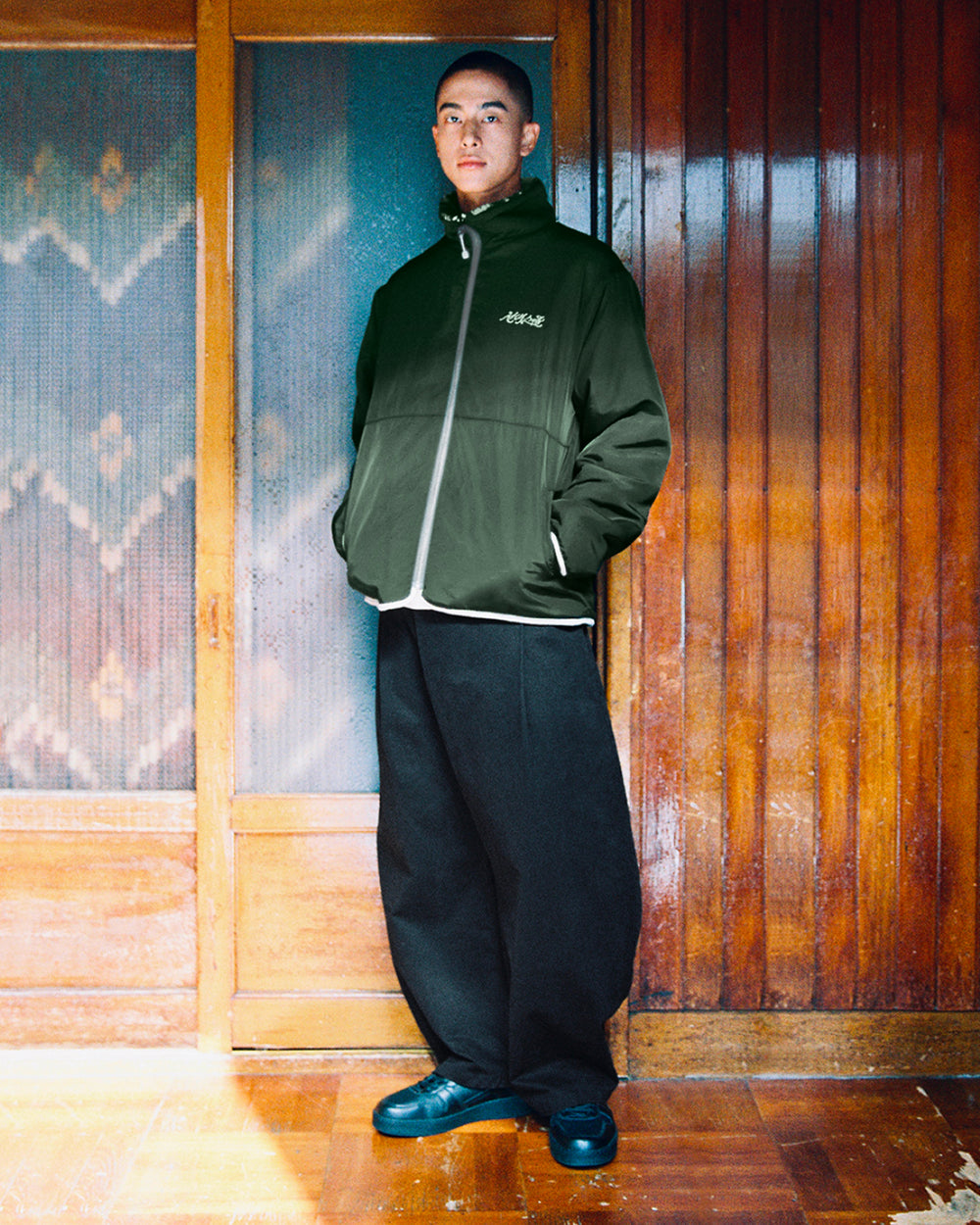 A person stands indoors in front of wooden doors, wearing the Sundae School TEAL REVERSIBLE GOSTOP FLEECE with a stand collar, paired with black wide-leg pants and black shoes, hands in pockets and a neutral expression.