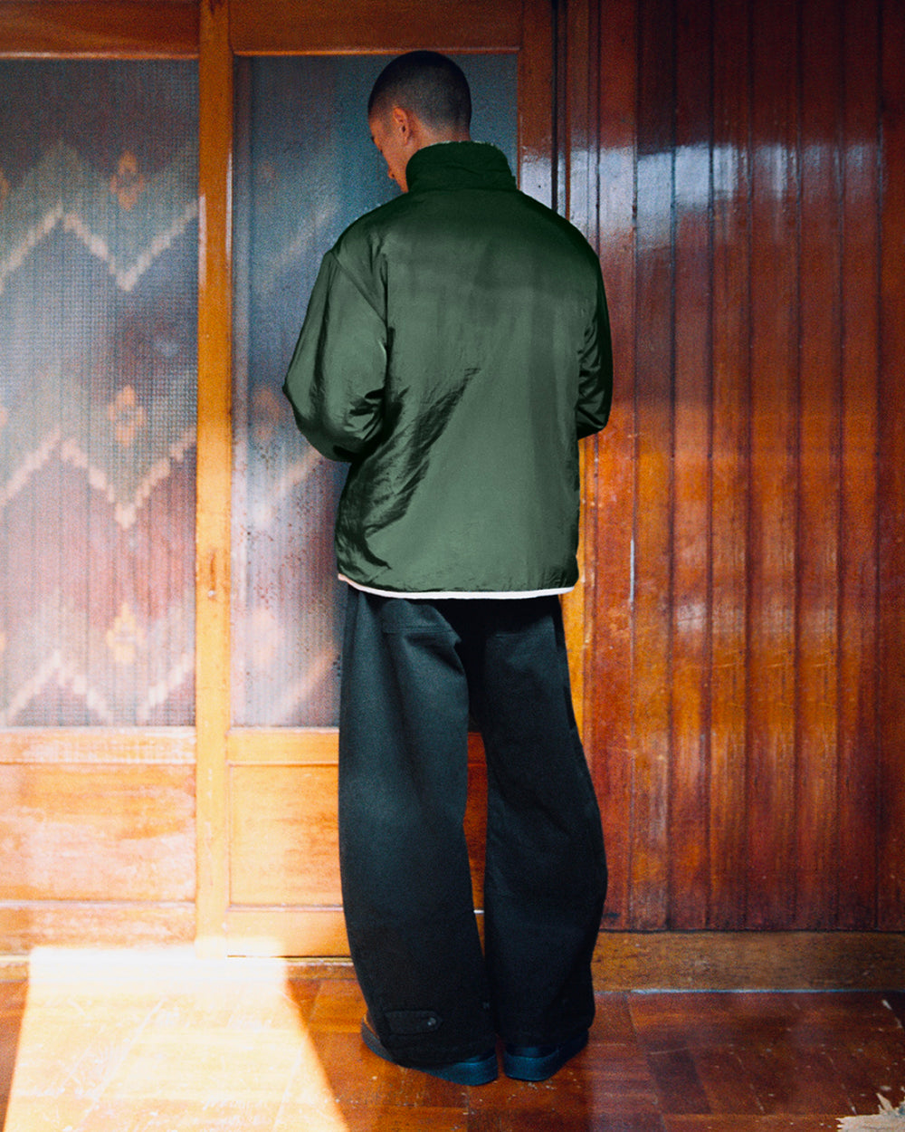 A person with short hair stands indoors facing wooden sliding doors, wearing the Sundae School TEAL REVERSIBLE GOSTOP FLEECE—a dark green sherpa jacket with a stand collar zip up—and very wide black pants.