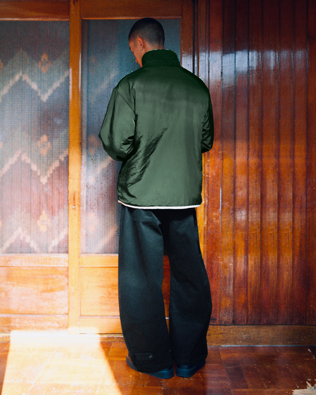 A person with short hair stands indoors facing wooden sliding doors, wearing the Sundae School TEAL REVERSIBLE GOSTOP FLEECE—a dark green sherpa jacket with a stand collar zip up—and very wide black pants.