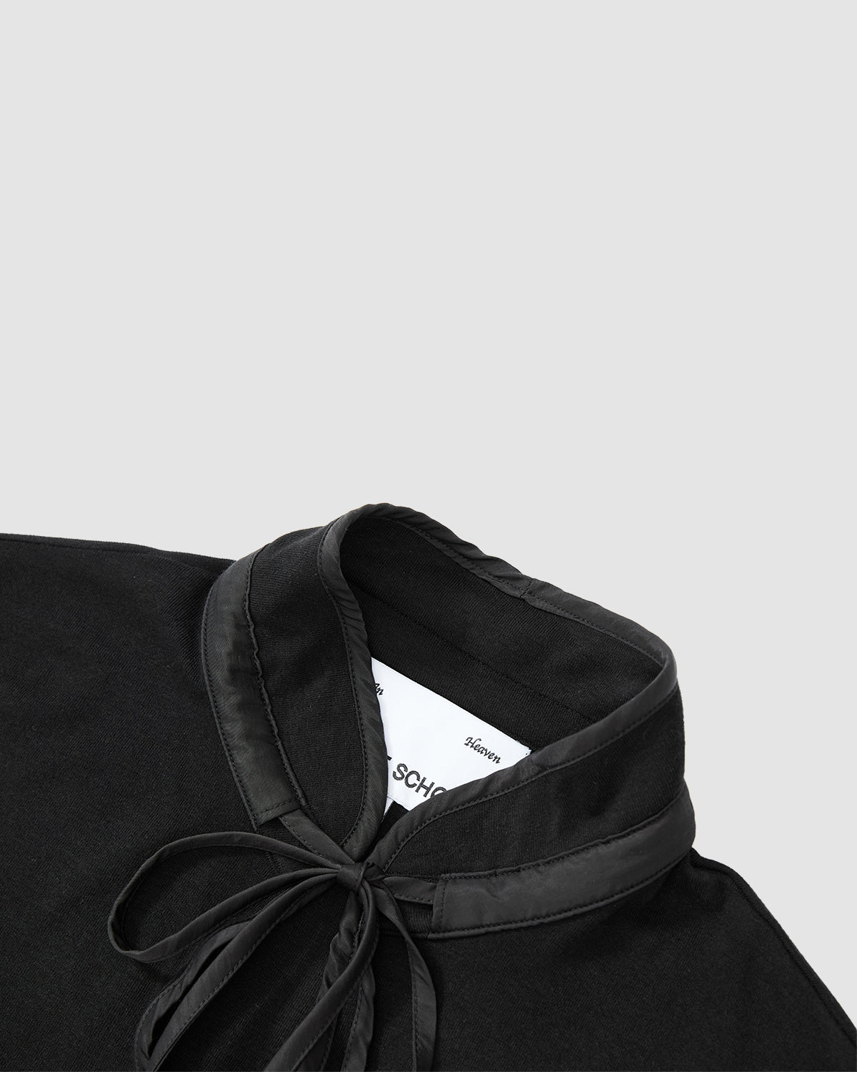 A close-up of the BLACK RIBBON TIE POLO by Sundae School features a satin-trimmed Manchurian teardrop collar and tied ribbon bow at the neck, set against a light gray background with a white label partly visible inside the collar.