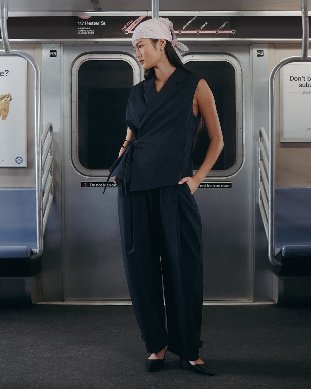 Wearing Sundae School's KIKI DRAWSTRING SLACKS, a woman stands alone in an empty subway car, gazing to the side amid station signs and posters.