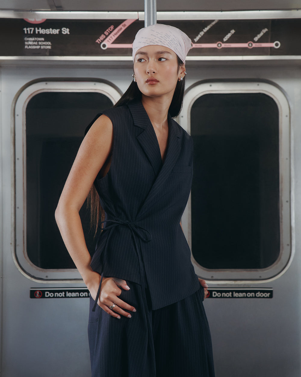 A woman in the Sundae School SABRINA VEST, a sleeveless pinstripe wrap top, and a light pink headscarf with a subtle Korean goreum tie detail stands calmly in front of “117 Hester St” subway doors, looking thoughtfully to the side.
