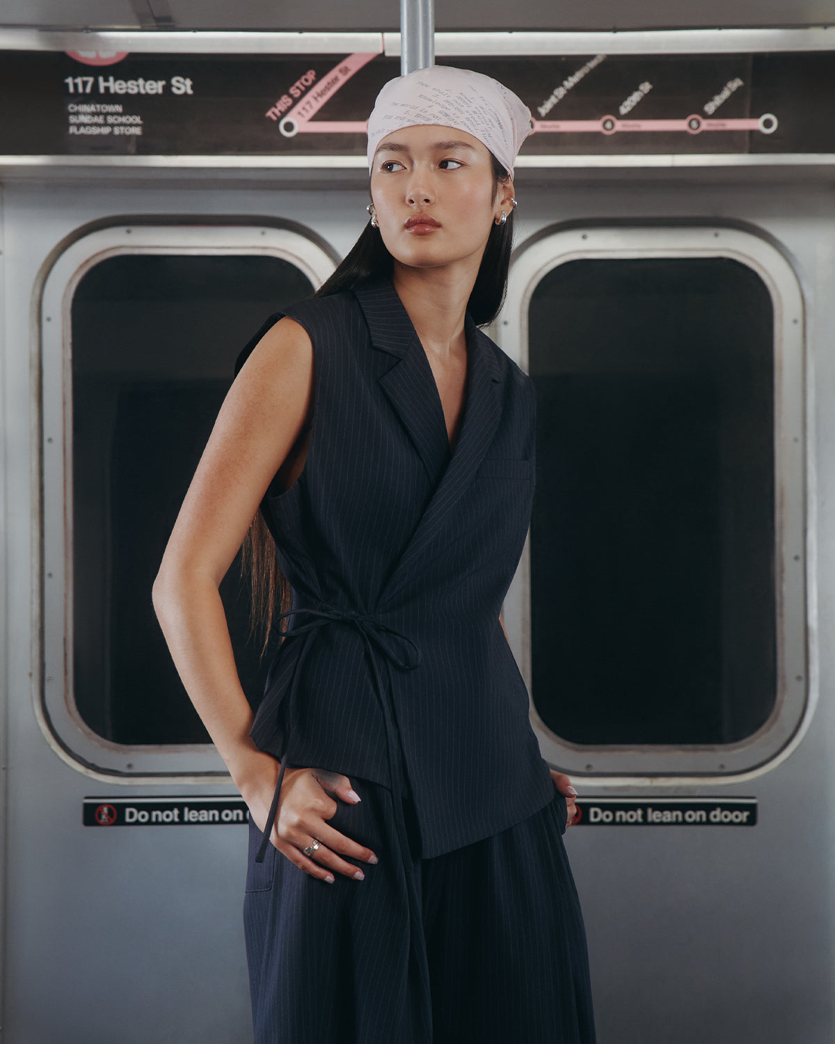 A woman in the Sundae School SABRINA VEST, a sleeveless pinstripe wrap top, and a light pink headscarf with a subtle Korean goreum tie detail stands calmly in front of “117 Hester St” subway doors, looking thoughtfully to the side.