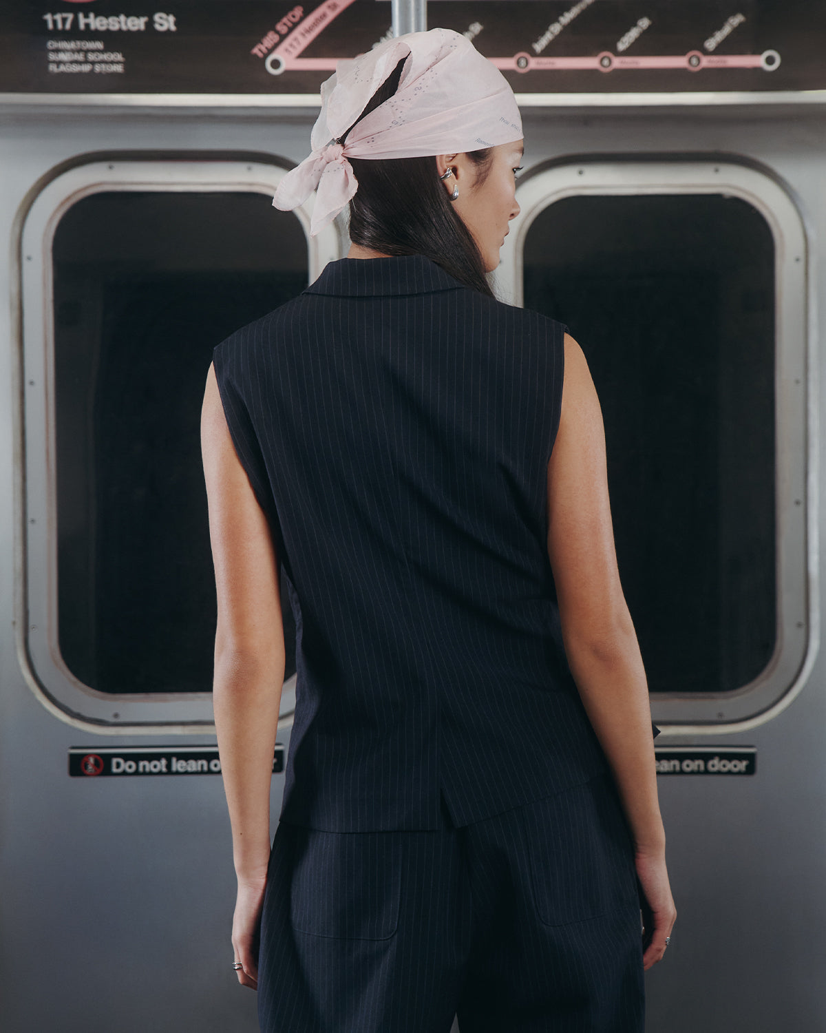 A woman wearing the SABRINA VEST by Sundae School stands with her back to the camera in a subway train, with signage and doors visible in the background.