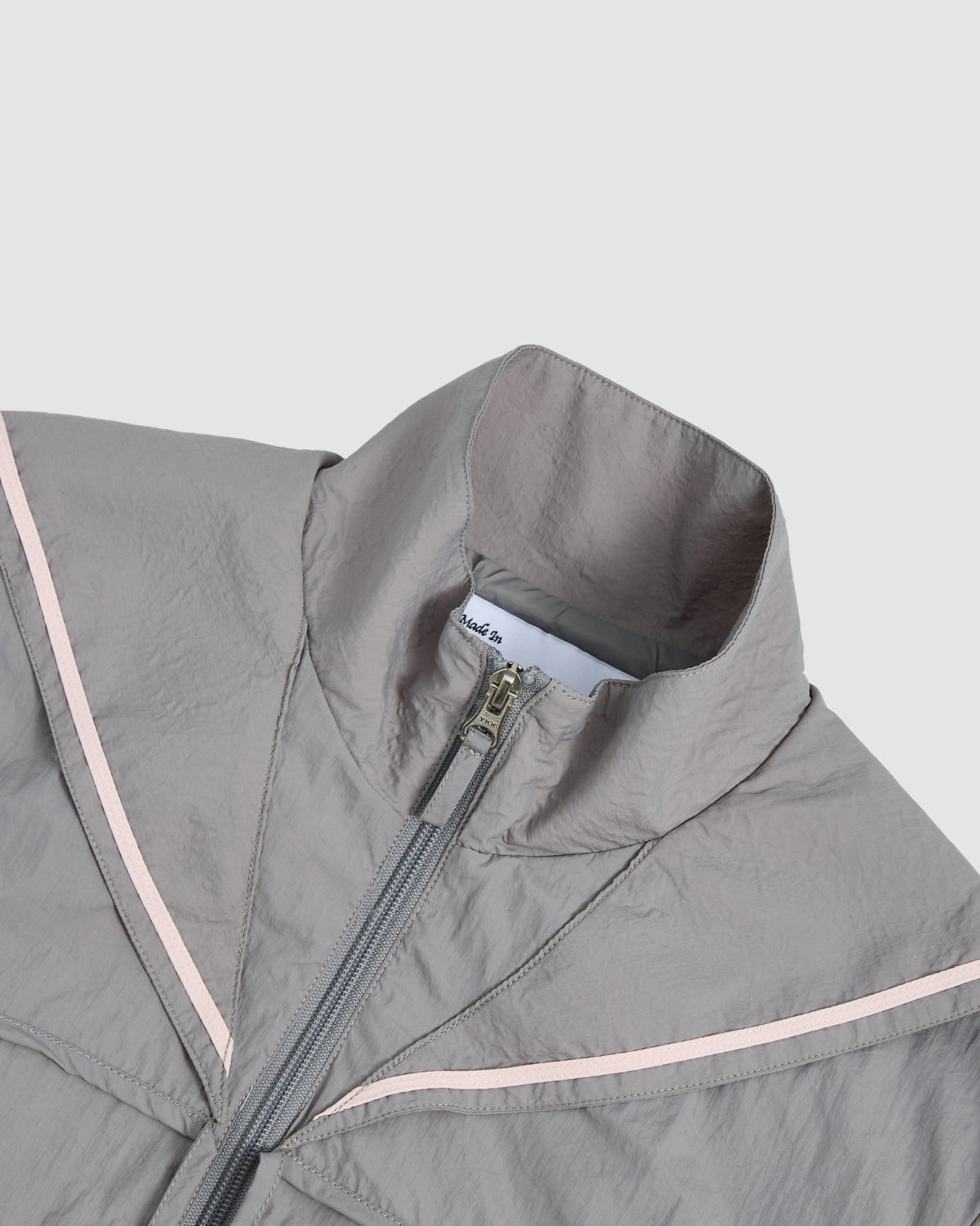 A close-up of the Sundae School CHARCOAL SAILOR WINDBREAKER in light gray, featuring a high collar, front zipper, and thin pale pink diagonal chest stripes, shown against a plain light background.